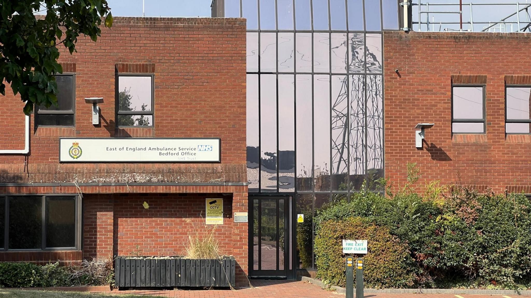 The Bedford office of the East of England Ambulance Service.  The building is shown close up with a white sign on the front and the trust logo, it is old red brick and has mirrored windows.