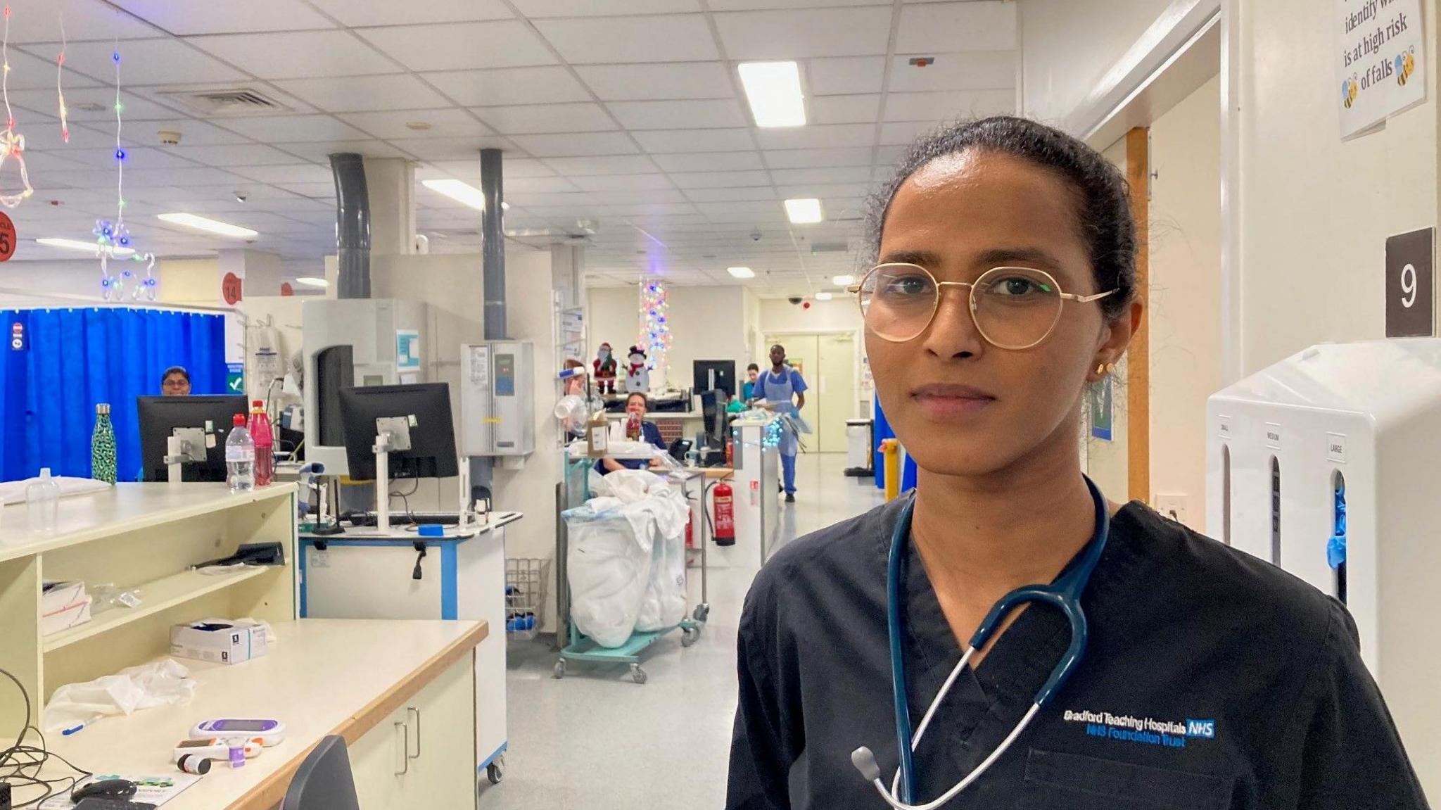 A woman with round, gold-rimmed glasses and hair scraped back into a ponytail with a stethoscope tied around her neck and wearing a dark blue medical tunic.