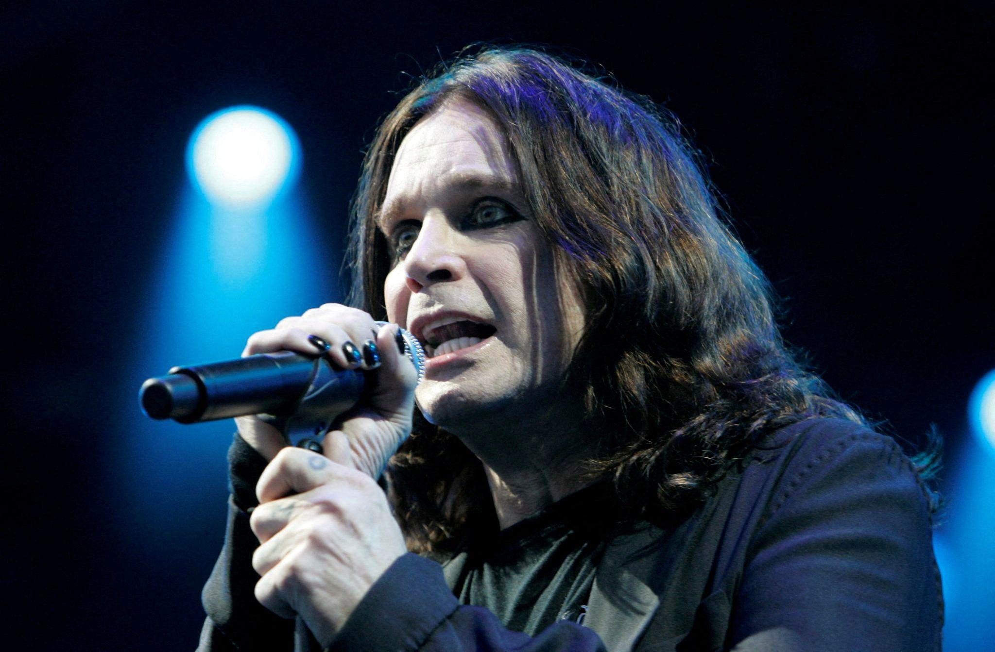 Ozzy Osbourne ka flokë të gjatë e të valëzuar të errët dhe mban një mikrofon të zi afër gojës ndërsa performon në skenë. Personi ka veshur një kostum të zi dhe thonjtë i ka të lyer me të zezë. Një dritë blu e ndritshme shkëlqen në sfond, duke krijuar një atmosferë dramatike koncerti me një mjedis të errët dhe ndriçim të fokusuar skenik.