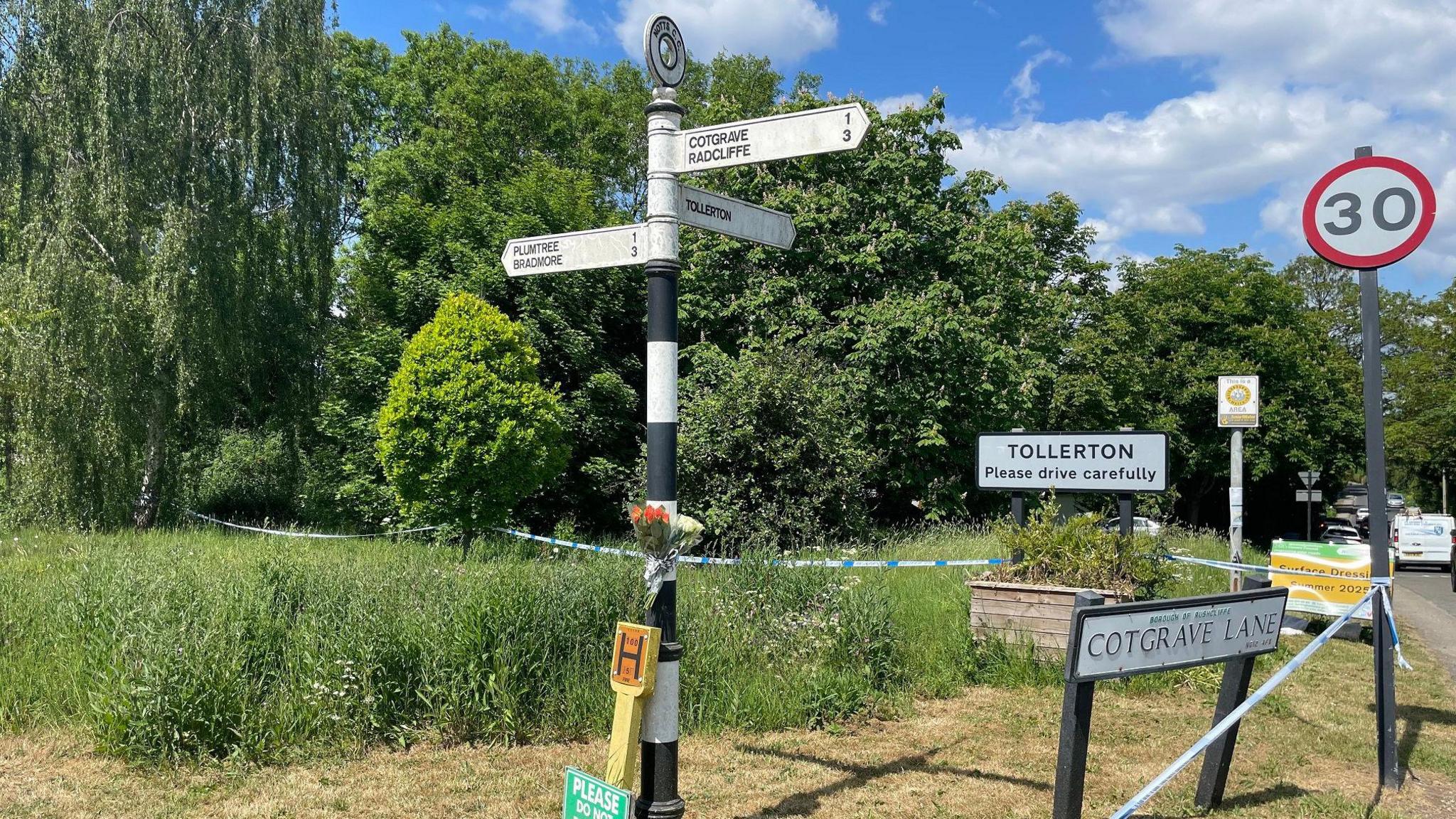 A bouquet strapped to a fingerpost sign with police tape nearby at the entrance to Tollerton village
