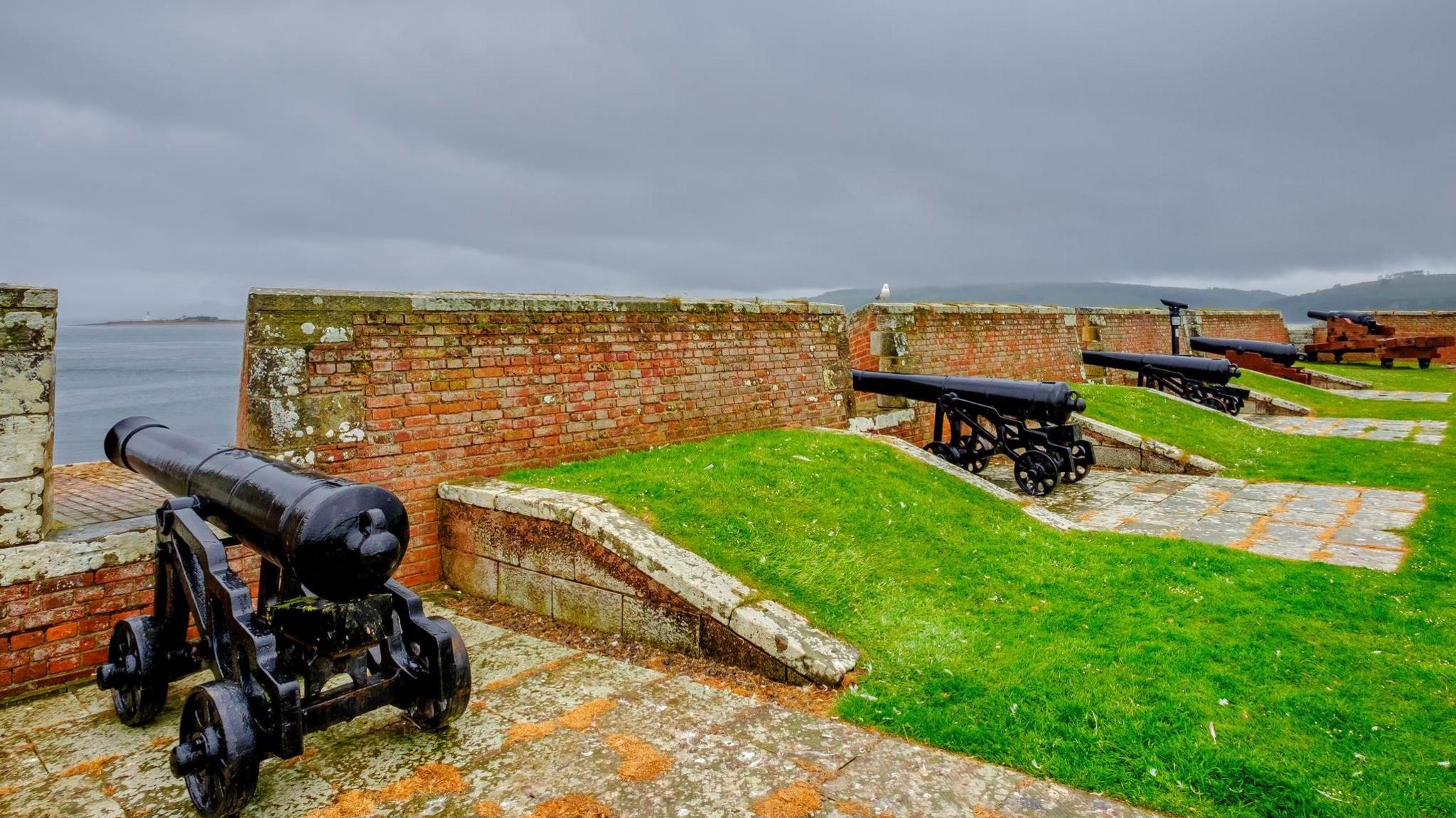 Cannons, which have been painted black, are lined up a long a wall of the fort. There are strips of grass between each gun.