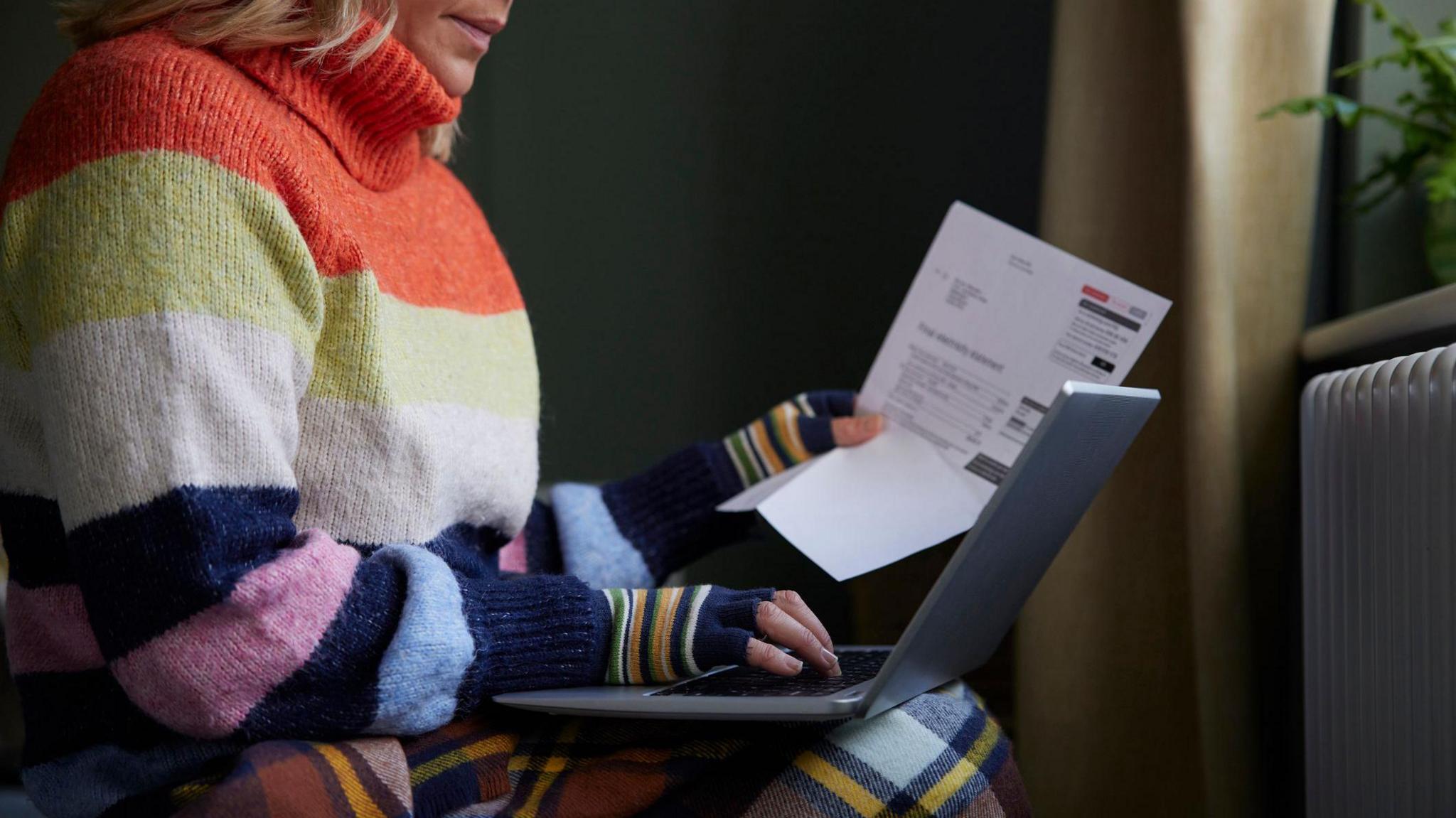 A woman in gloves with laptop and bill trying to keep warm by radiator during cost of living energy crisis