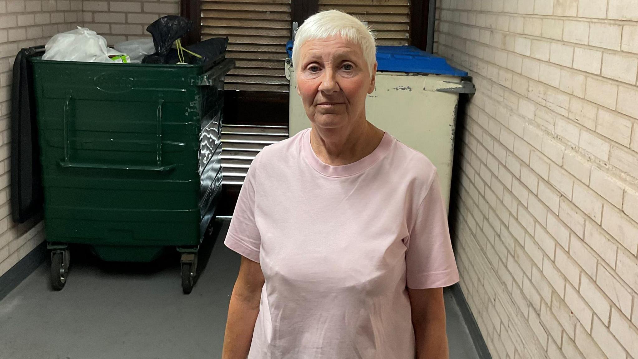 A woman with short white hair and wearing a pink t-shirt standing in front of two large bins. One has some white and black bags in and the other is closed.