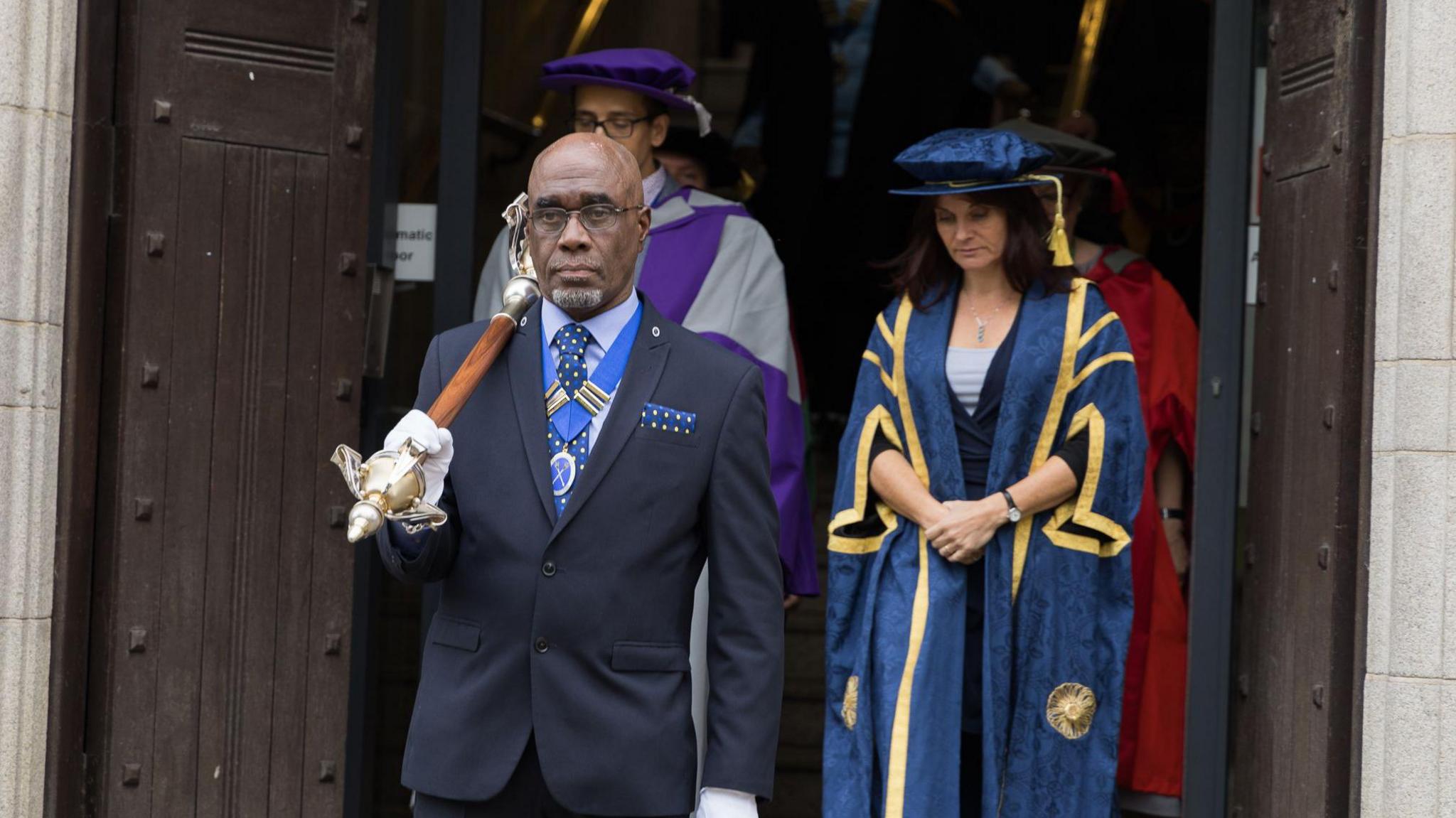 David Bourne wearing a black blazer, glasses and a blue spotted tie. He is holding a mace whilst wearing white gloves. Behind him, a woman is wearing a blue and gold gown and a man stands in a purple and grey gown