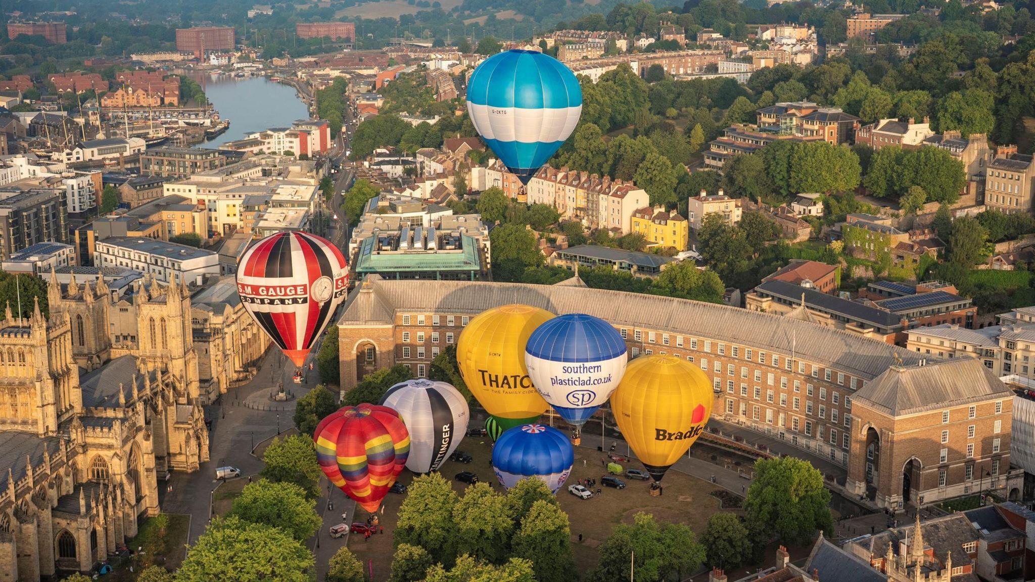 A group of colourful hot air balloons are seen from a drone shot, taking off from College Green in Bristol. City Hall is clearly visible as is the waterways of the floating harbour and much of Clifton