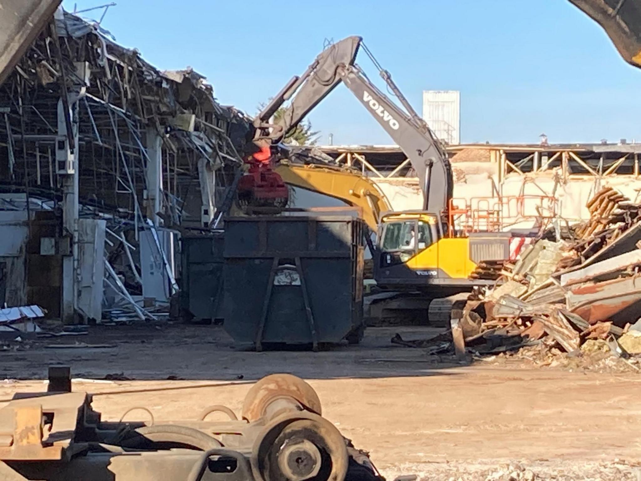 A digger is working on demolishing the former Delphi building. Parts of the building have been torn down with twisted metal visible and debris on the floor