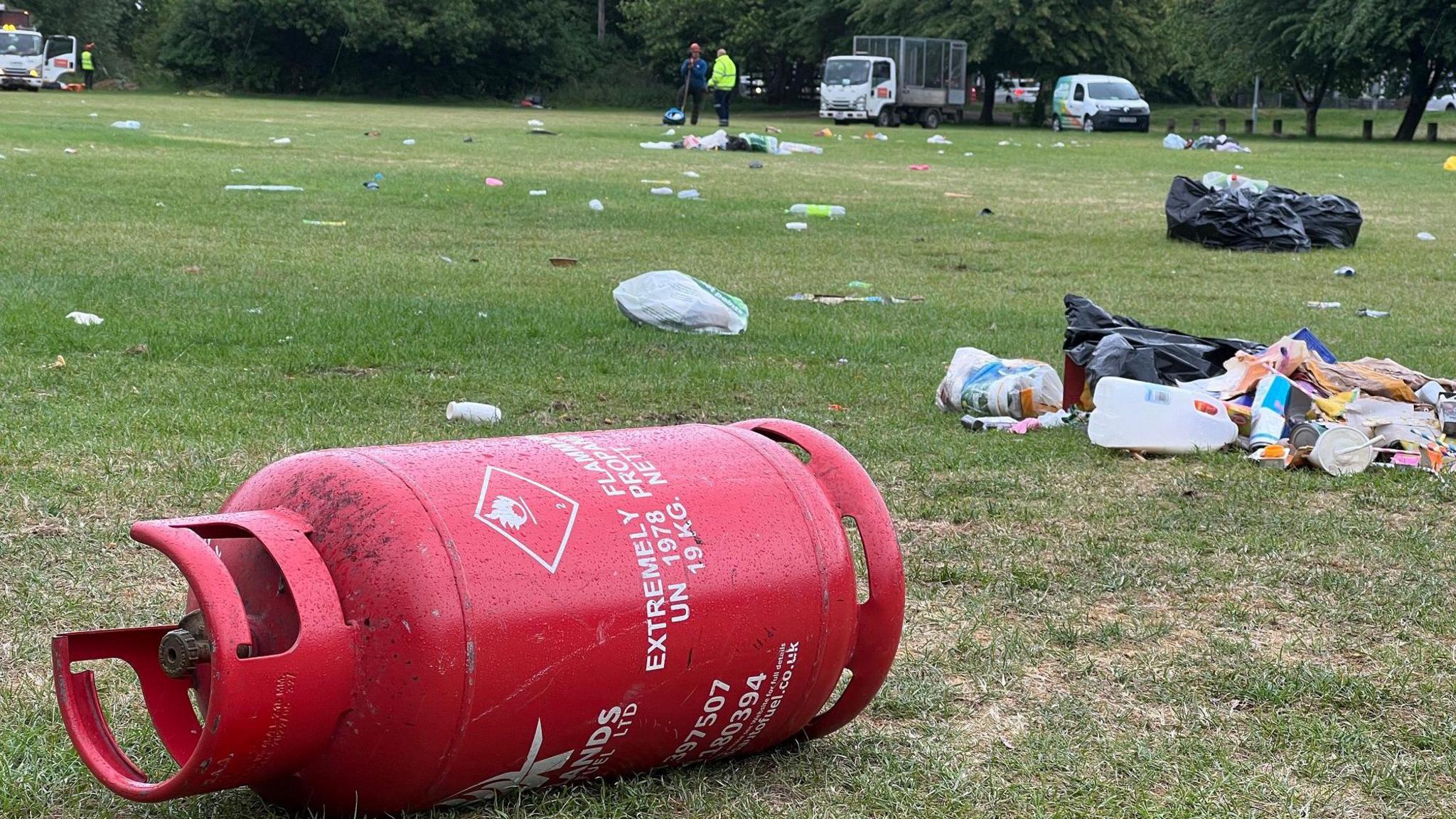 In the fore of the photo is a large red gas canister with piles of household rubbish scattered in a green field behind