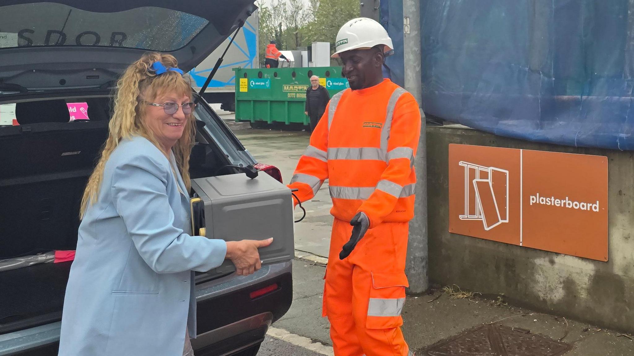 Woman with long light brown curly hair wearing a light blue coat and carrying a grey object which she is handing to a recycling operative in an orange hi-vis and white hard hat. There is a grey car with its boot open behind the woman and a skip covered in a blue tarpaulin behind the recycling operative. It has a red sign which says "plasterboard" in white lettering.