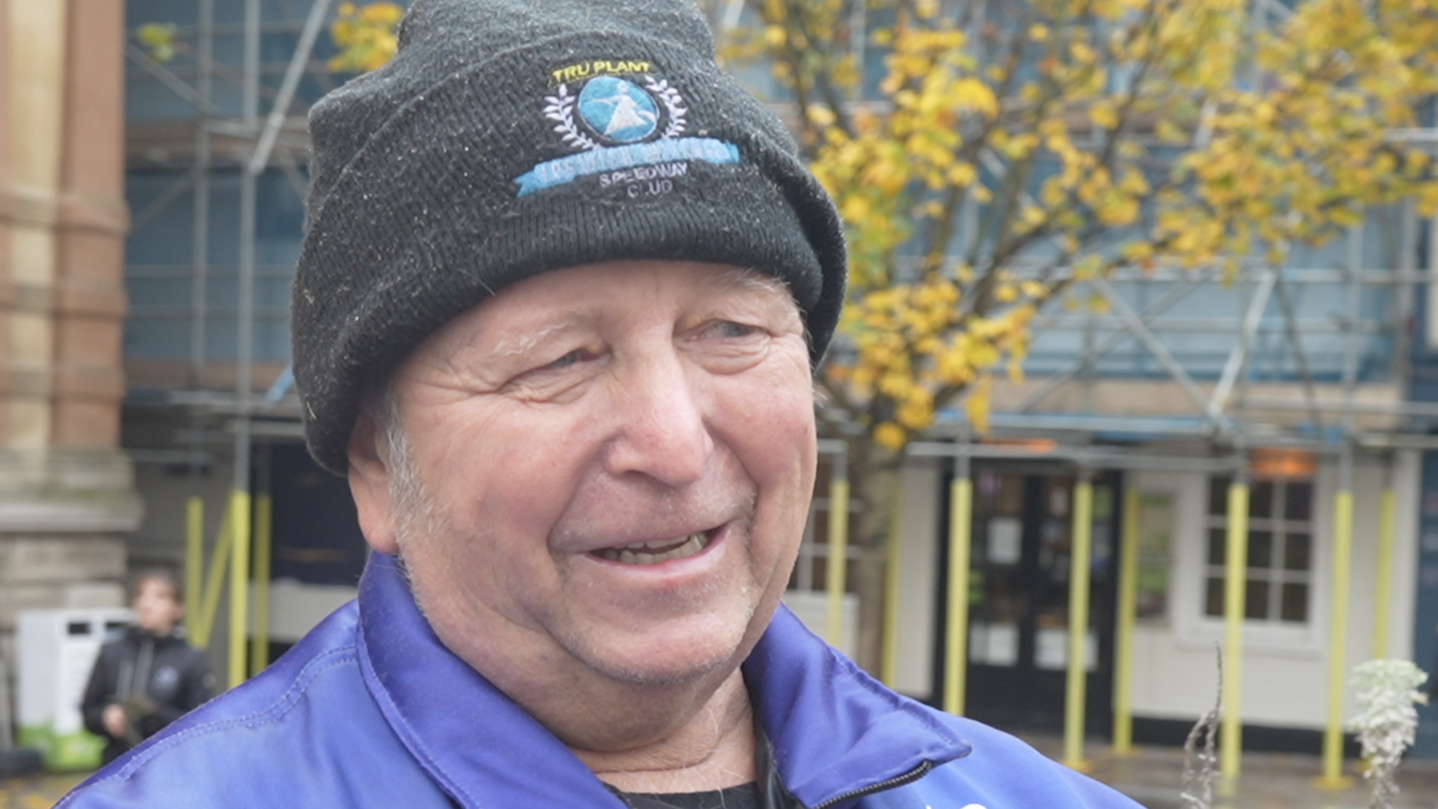 Julian Moore looks away to the right as he talks. He wears a grey beanie hat and blue jacket. A tree and a building with scaffolding around it can be seen behind him.