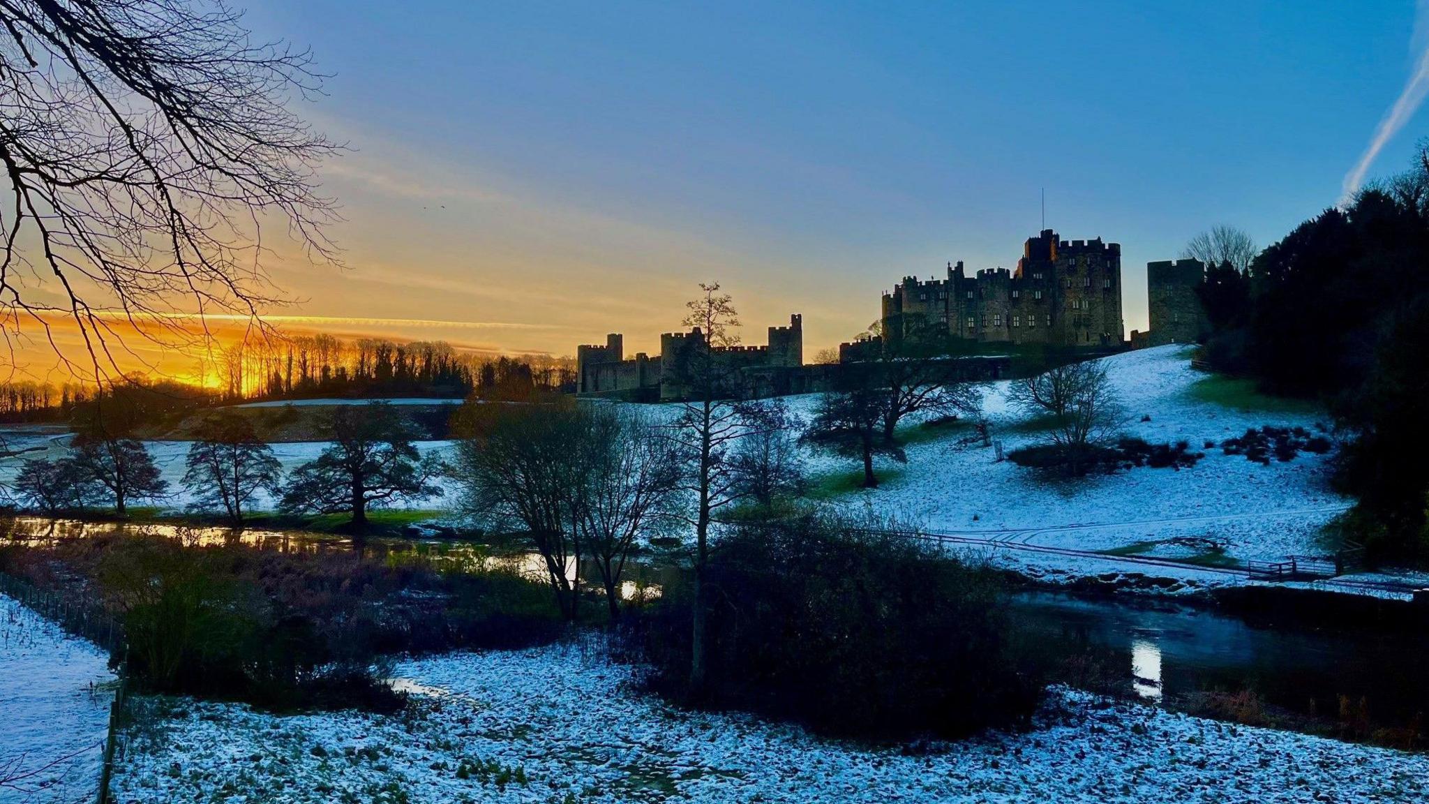The sky turns orange as the sun is about to rise at Alnwick Castle, a medieval stone structure with towers and windows. The land around the castle is covered in a thin layer of snow.