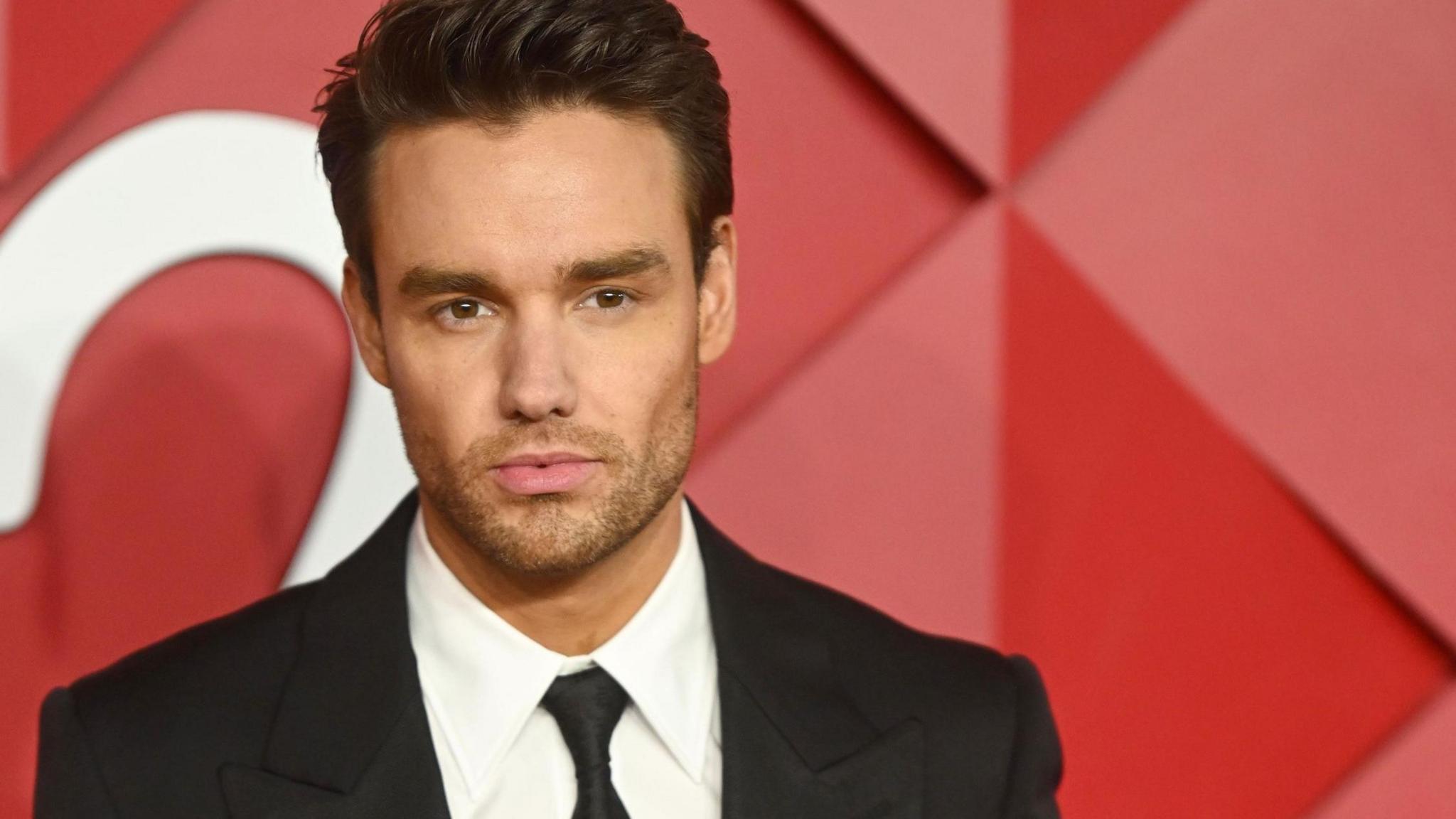 Liam Payne, wearing a black tuxedo, poses against a red background