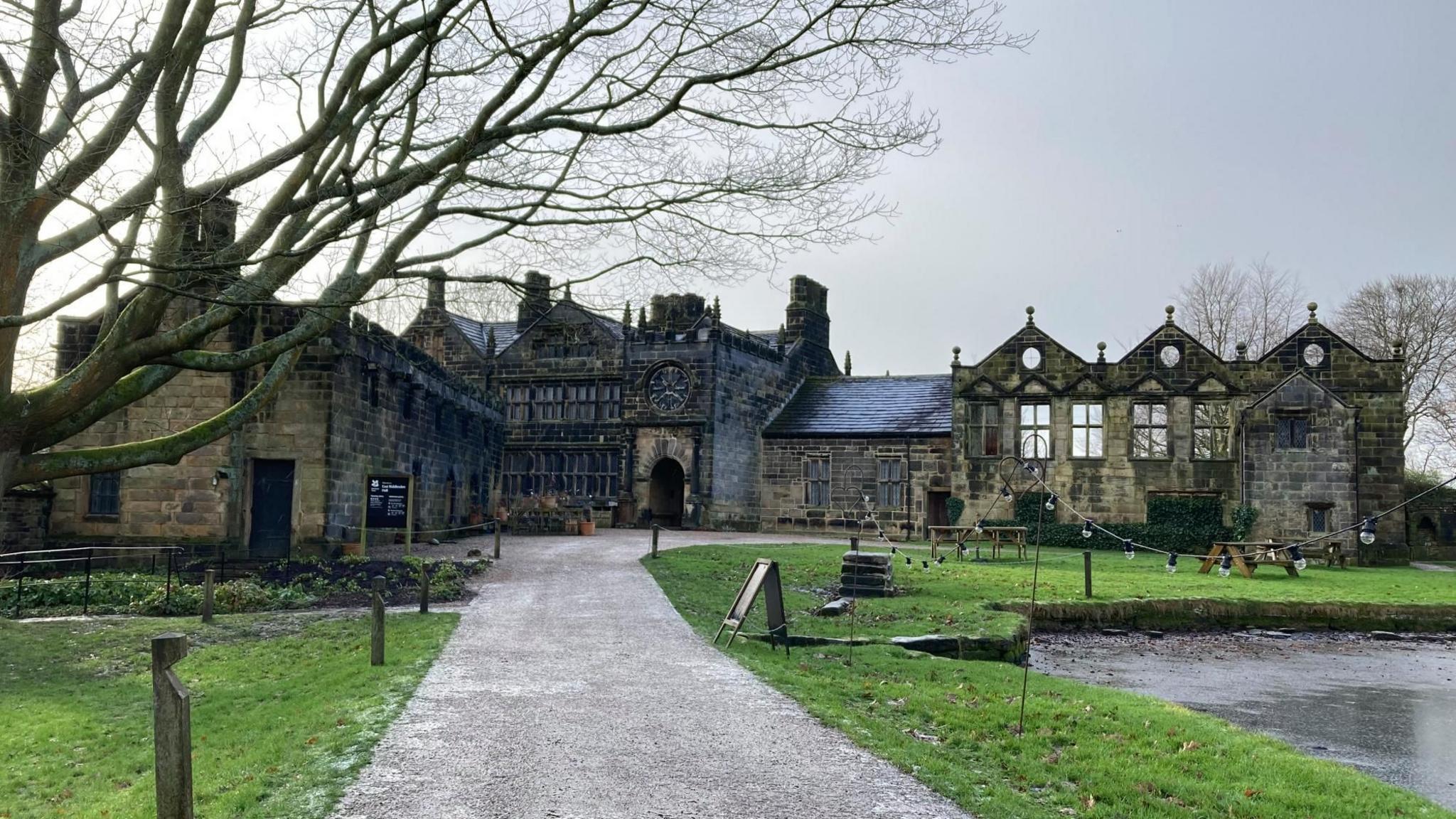 A partially ruined stone manor house, set in grassy surroundings with a pond to the right and a tree to the left and a gravel path leading up to its entrance