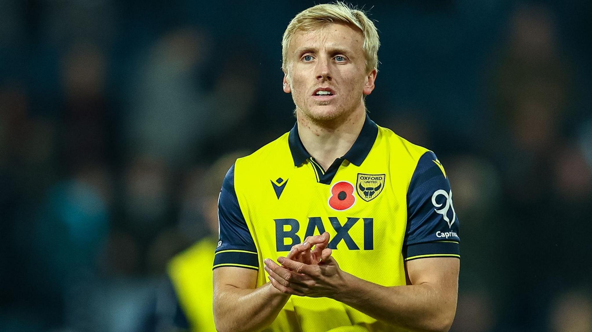 Louie Sibley, wearing the Oxford United yellow and navy home shirt with a poppy on the chest, applauds the U's fans