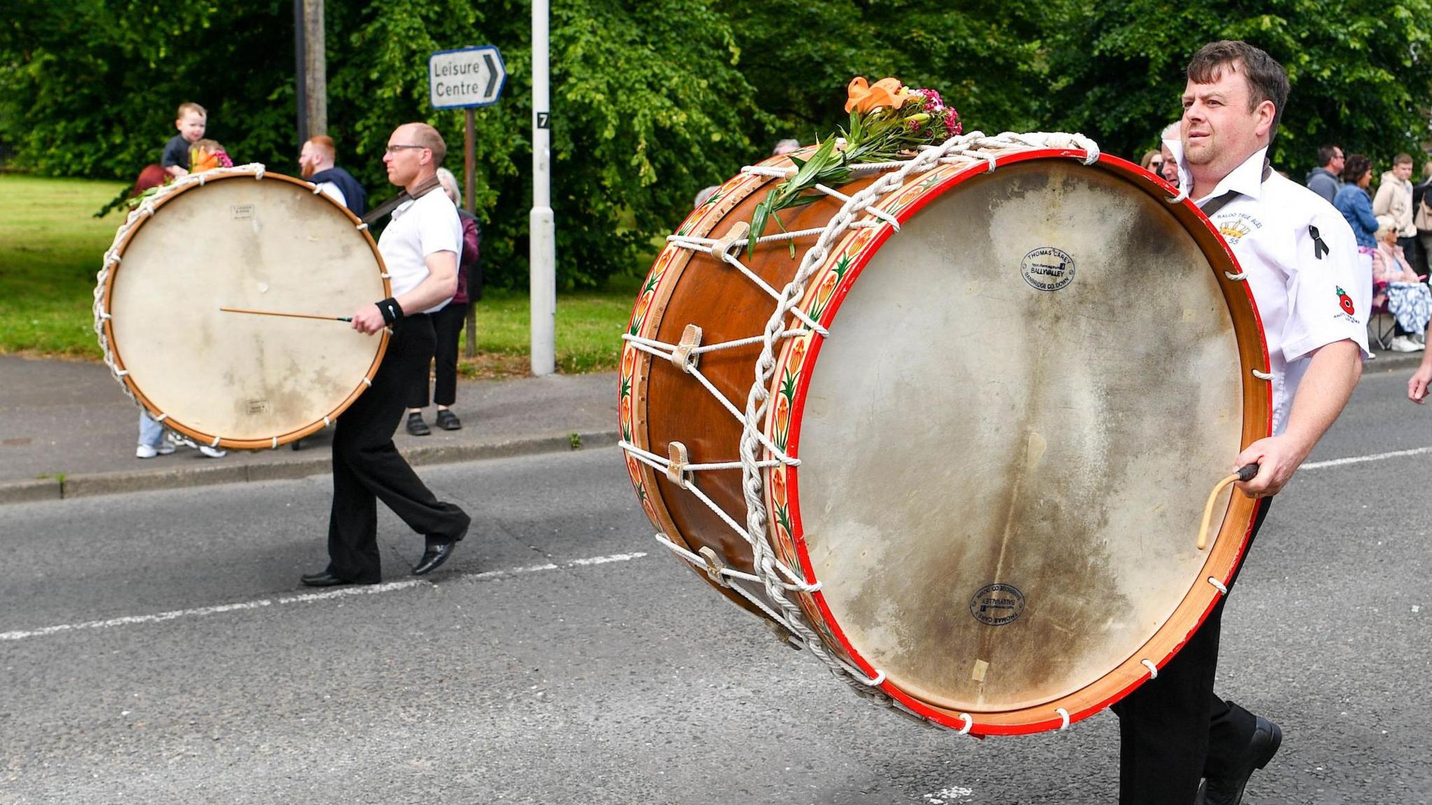Two band members playing Lambeg drums during a 12th July parade in Carrickfergus last year. The huge, wooden, double-sided drums are worn on straps across the front of their bodies and they stretch in length from their face to their knees. The drummers are both wearing white shirts and black trousers.