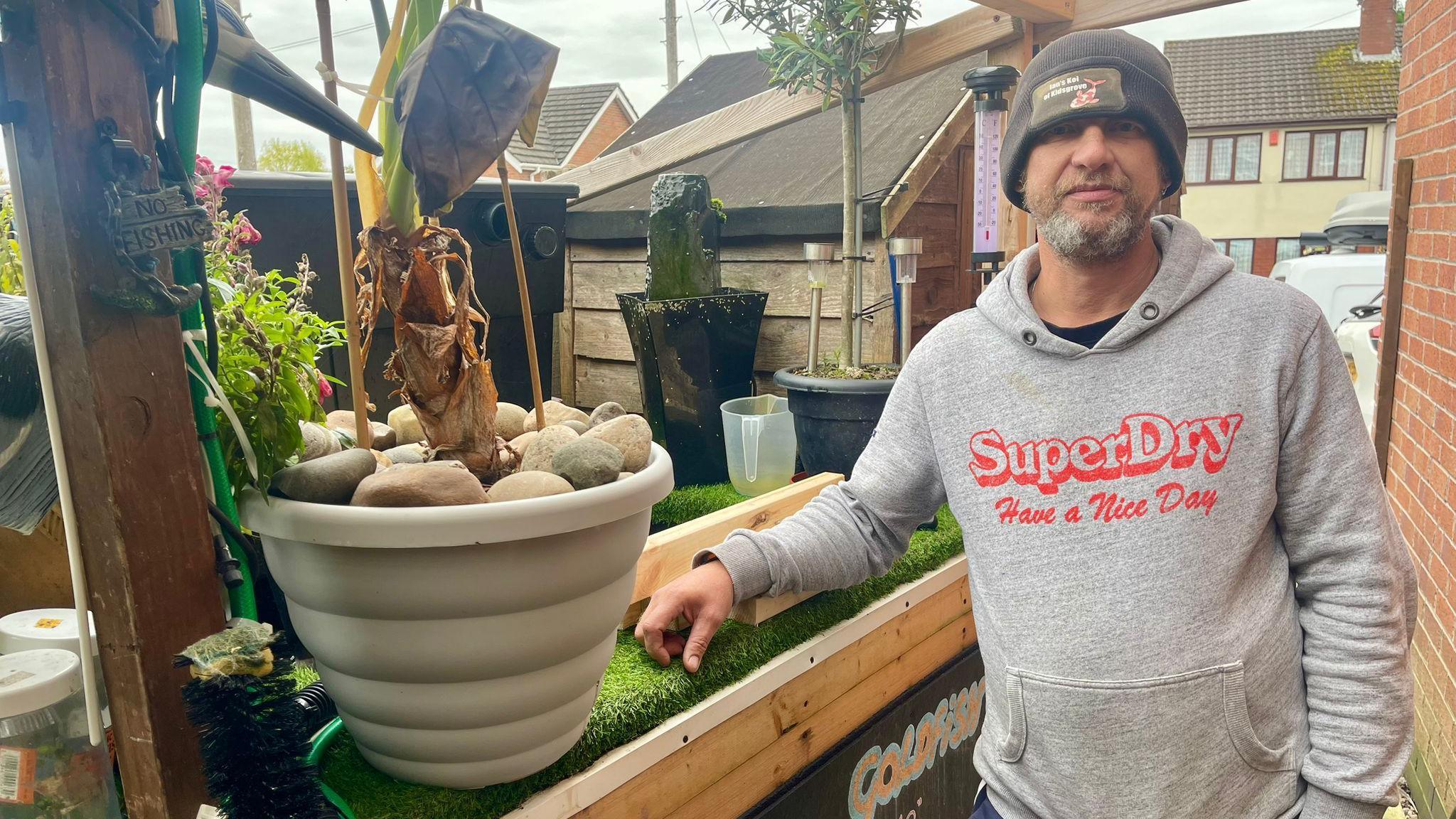  A man is a black woolly hat and grey hoodie stands beside an artificial pond. He has a grey beard and rests his arm on a planks of wood. There is a large plant pot full of stones and an exotic plant. Other plants and a waterfall can be seen. A shed and the wall of a house can been seen close in the background.