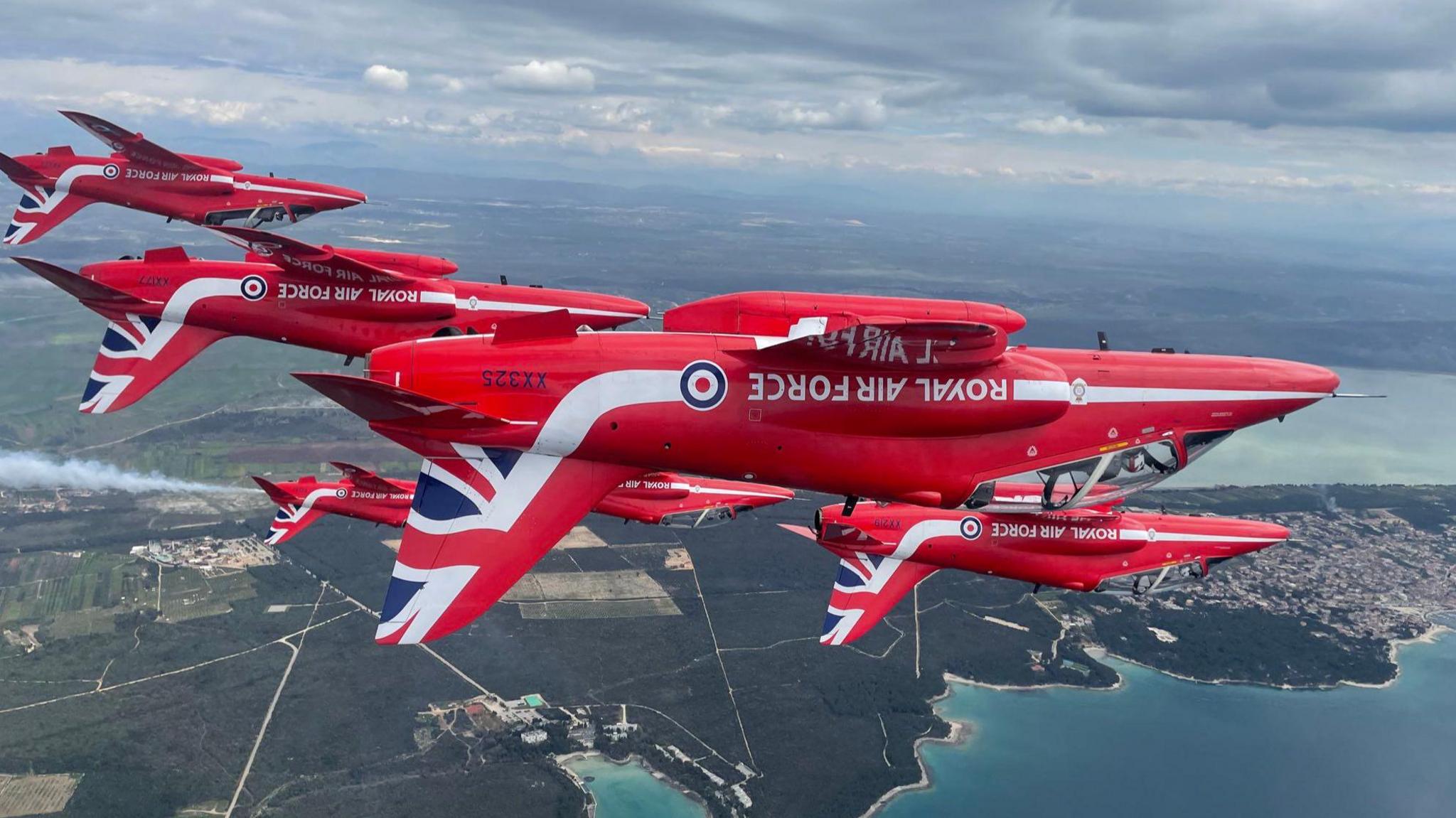 Red Arrows bearing Union flag livery flying upside down in formation along a coastal strip with land in the background