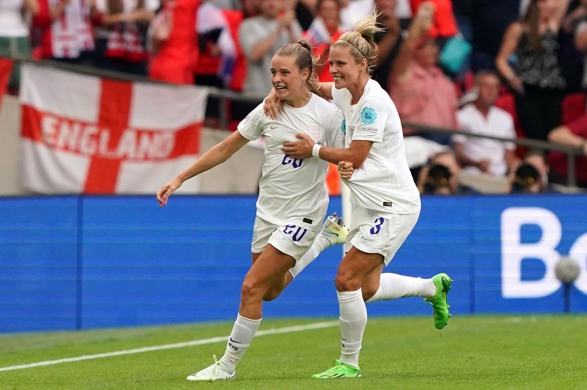 Two football players, Ella Toone and Rachel Daly, dressed in the white England strip, run towards the stands to celebrate with the fans after Toone scored at the Women's Euros in 2022. They look both happy and overcome with emotion.
