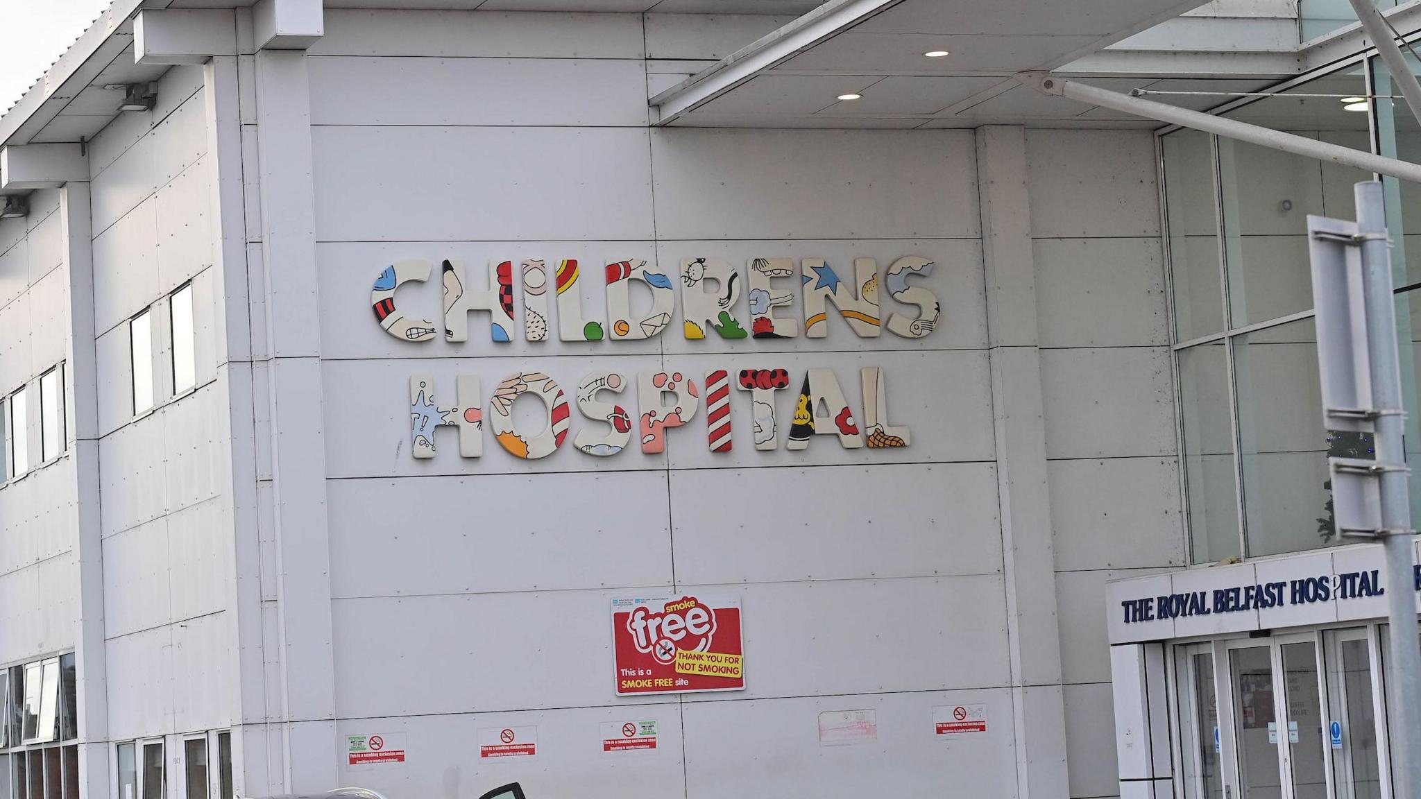 The exterior of the Royal Belfast Hospital for Sick Children. The outside walls are white and modern-looking and have the words 'Children's Hospital' in large colourful letters on the wall near the front entrance. The roof is green and above the doors is a section that is all glass panelling.