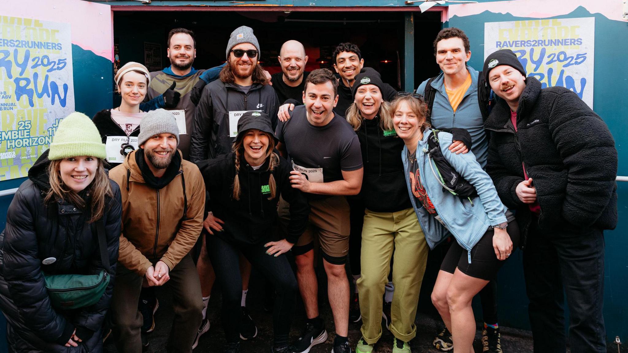 A group of people who are part of Ravers to Runners smile at the camera as they pose for a group shot. Some of them are in running gear and some are also wearing bobble hats