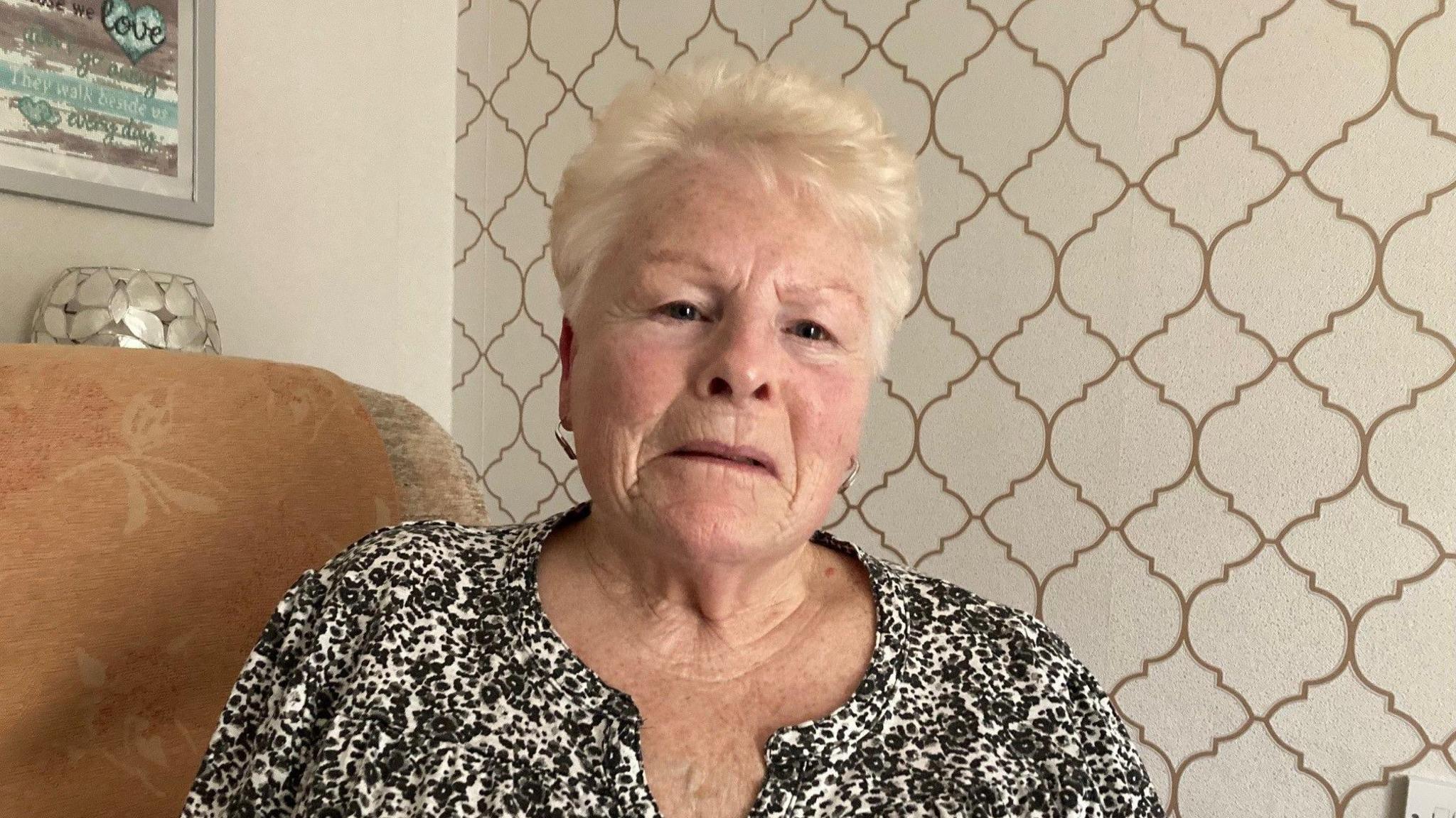 An elderly woman with dyed-blonde, short hair wearing a black and white blouse sitting on a sofa. 