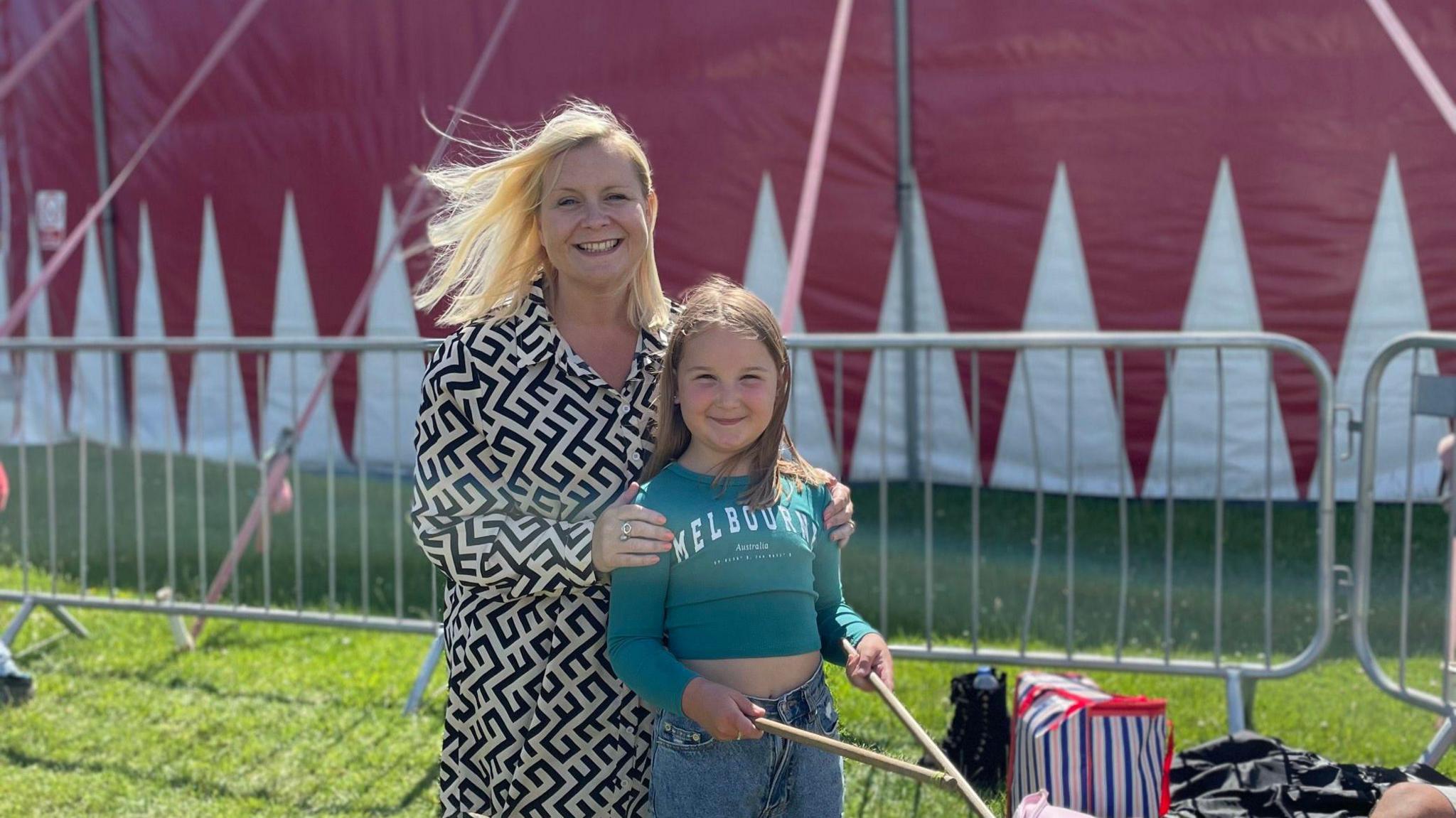 Nicola wears a white and black patterned dress with her hands on her daughter's shoulders. Lola wears a blue crop top and jeans and is holding wooden drumsticks. They stand in front of a red and white circus tent that has metal barriers in front of it. 