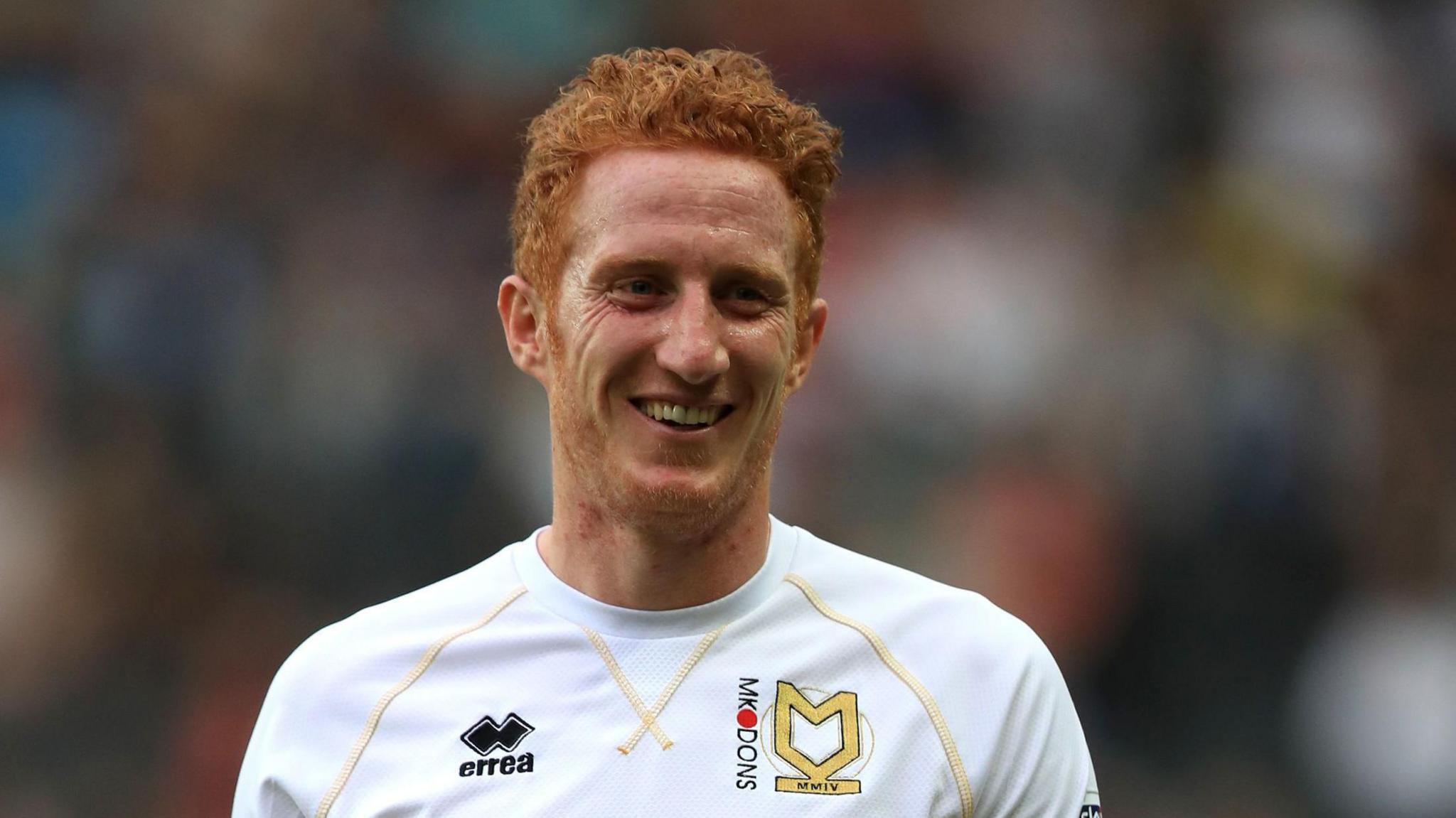 Dean Lewington is on a football pitch walking towards the camera. He has short ginger hair and a short bead. He is wearing a white football shirt with the MK Dons insignia on it.