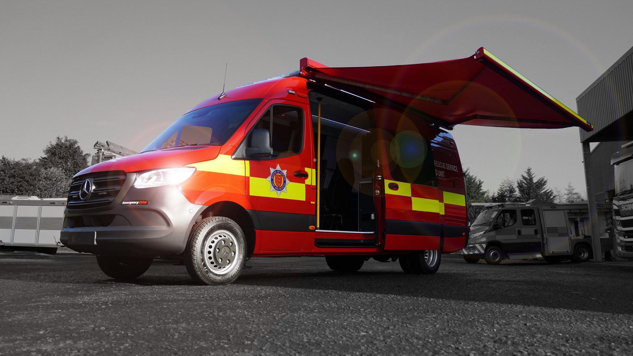 A red fire van with its sliding door open, revealing seats and communications equipment in the back of the van as well as an awning on the side, creating a covered area.
