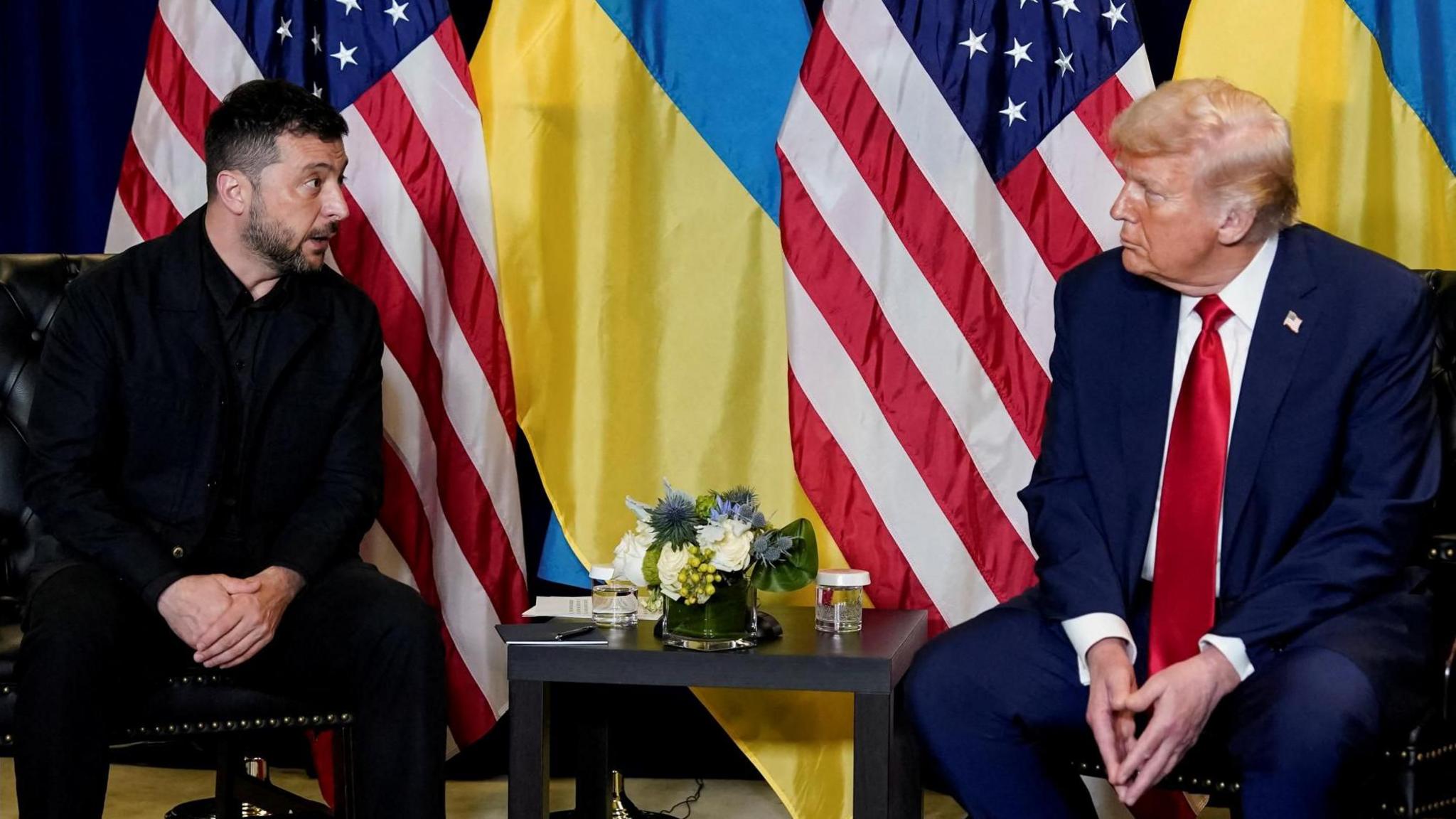 Zelensky, in dark clothing, and Trump, wearing a navy blue suit and red tie, sit in front of Ukrainian and US flags.