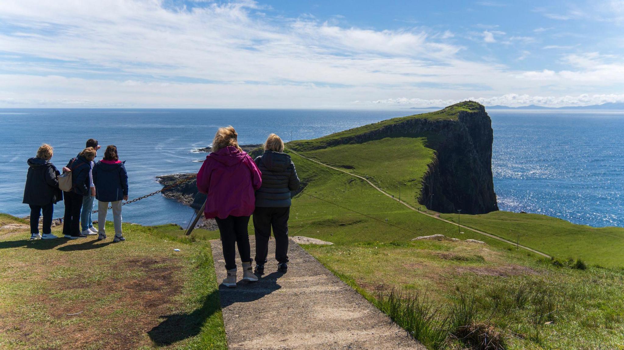Sianar dhaoine a' coimhead air rubha uaine agus a' mhuir. Tha ceum-coise a' sìneadh a-mach dhan rubha.