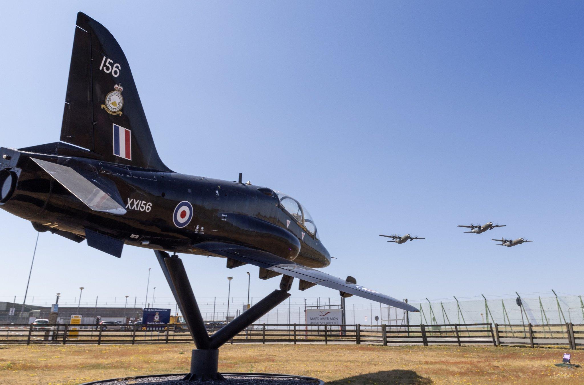 Three Hercules flying over a black Hawk aircraft on the ground at RAF Valley