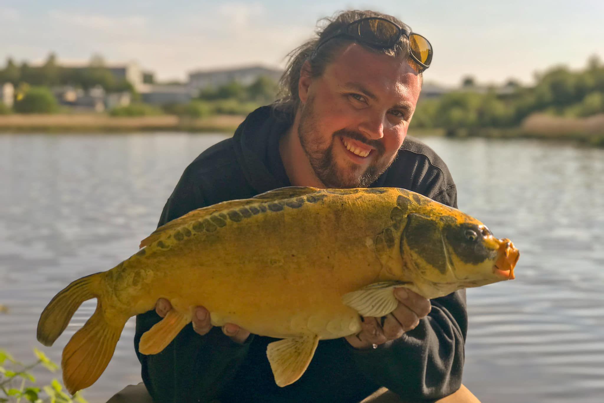 James Duffy smiling by a lake. He is crouched down holding a big carp fish. It is sunny and he has sunglasses on his head.