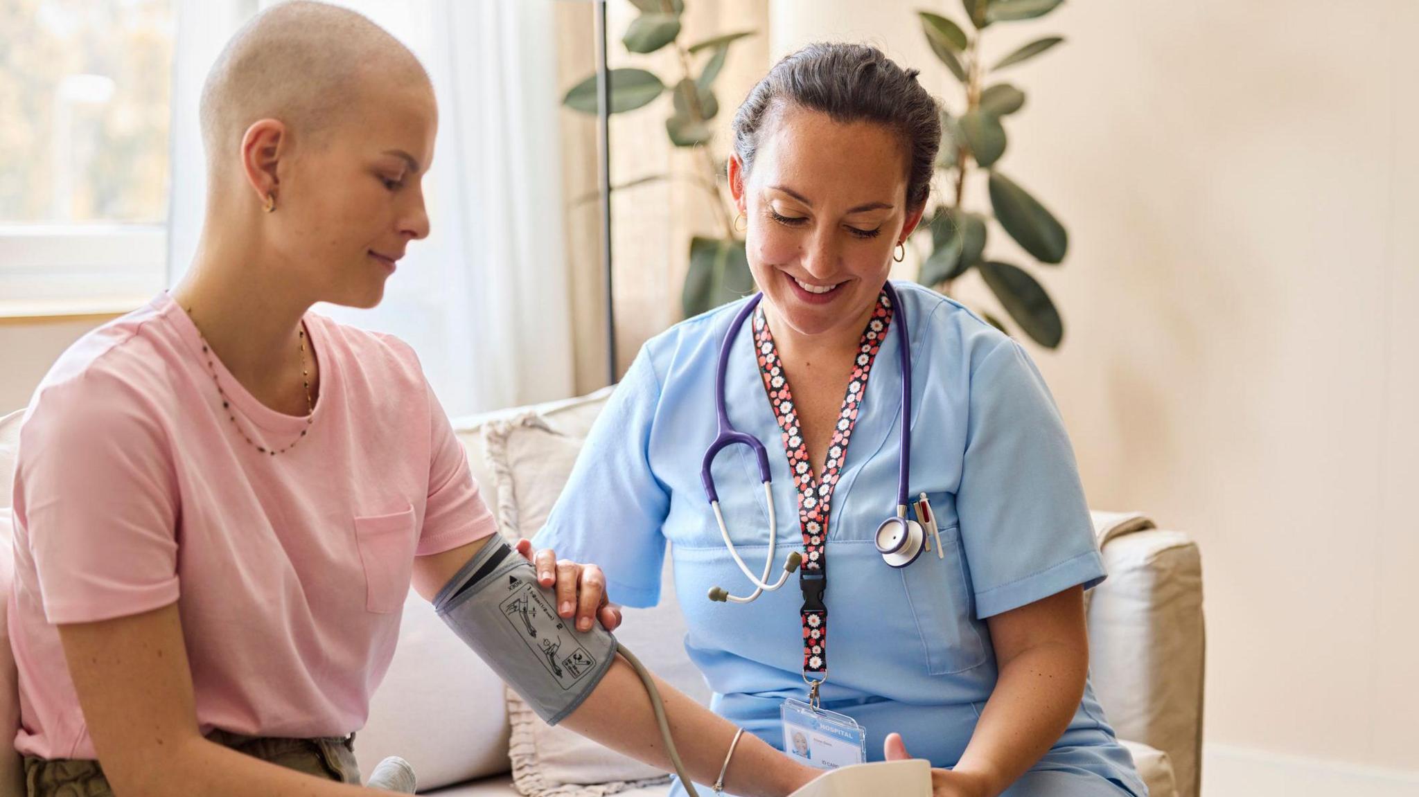Smiling female healthcare worker checking blood pressure of woman. Nurse wearing scrubs is examining patient at home. They are sitting on sofa.
