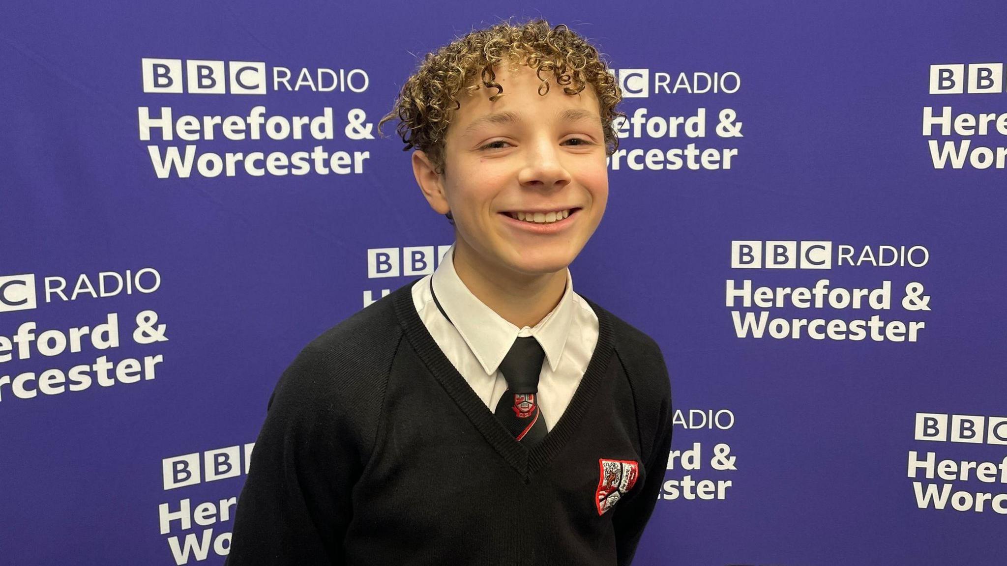 A teenage boy with curly hair stands in front of a purple backdrop decorated with the BBC Hereford & Worcester logo. He is wearing a school uniform - a white shirt with a black tie and a black sweatshirt with the Chantry School logo on the front.