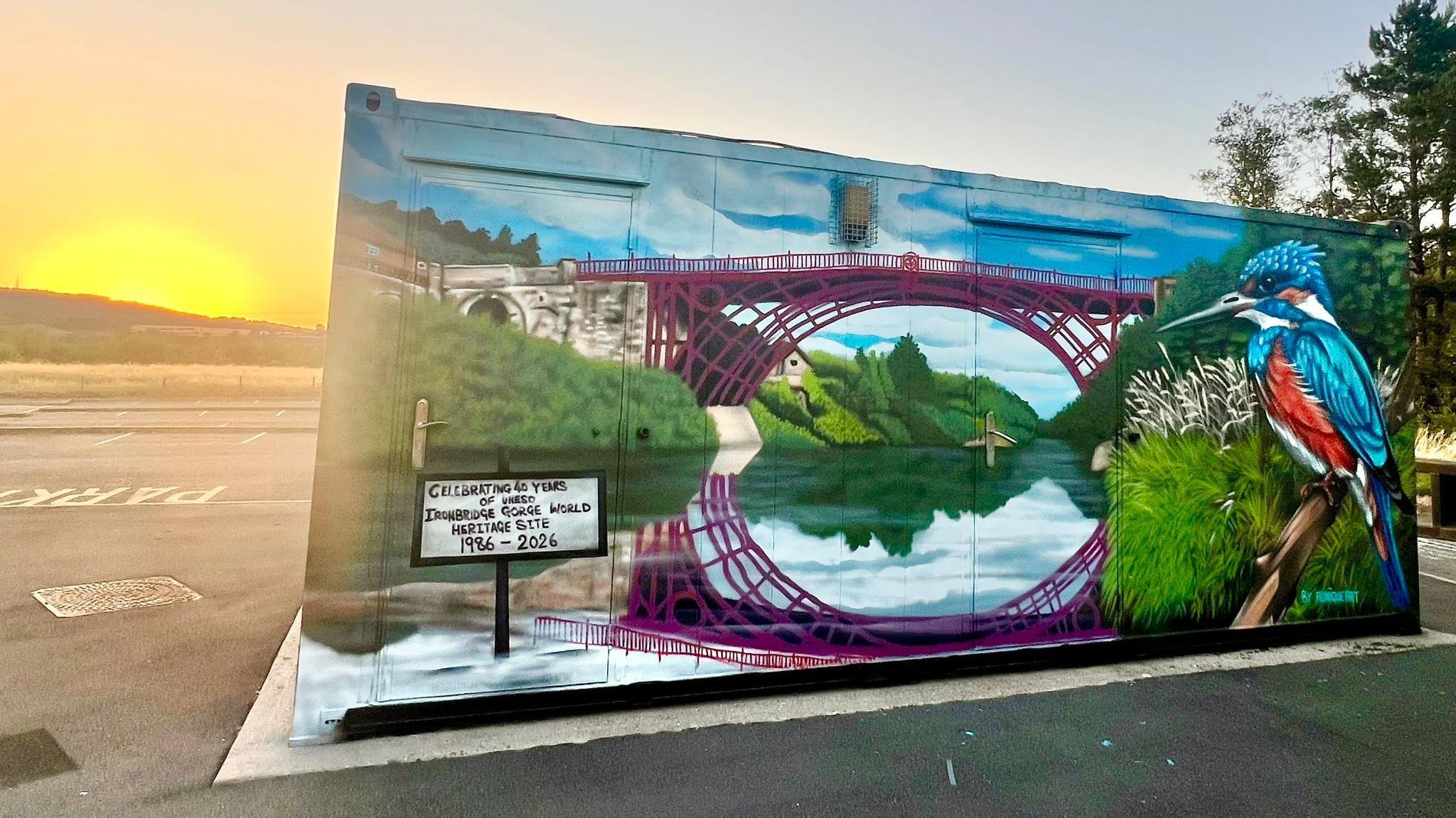 A graffitied hut of an iron bridge with grassy banks over a river. There is also a painting of a large kingfisher. The hut is on a car park with the sun setting behind a hill in the background 