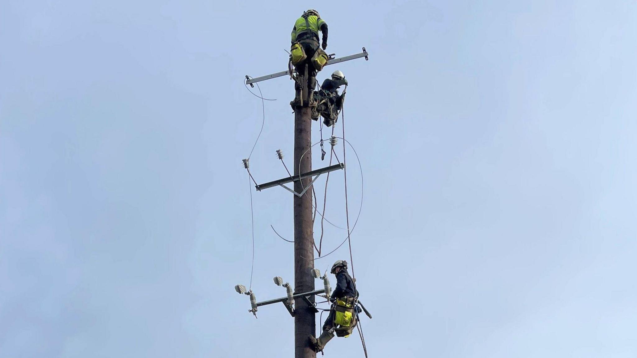 A wide shot of an electricity pole against a grey-blue sky, three workers in high-viz and helmets are working on it