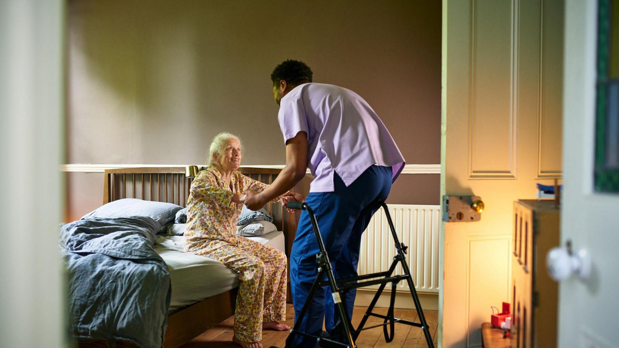 A care home nurse in a light purple tunic and blue trousers helping an elderly woman in brown pyjamas out of bed. There is a walker positioned nearby and the door is open with the light from a lamp reflected on the door, giving a warm glow.