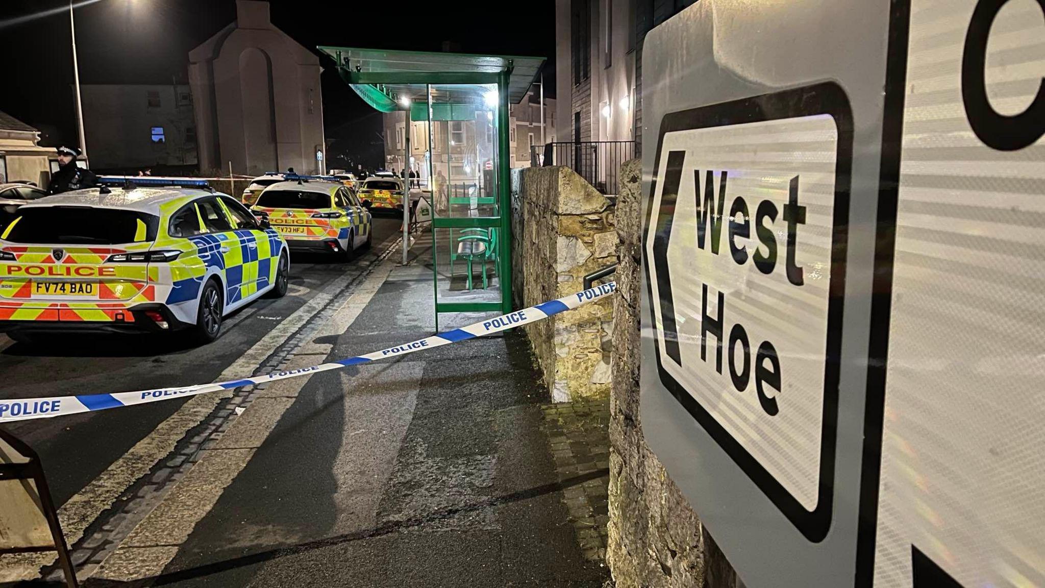 A West Hoe sign on the right with police tape across a pathway. There are several police vehicles behind the police tape, along with a bus stop on the pathway. The photo was taken at night so the pathway and road is lit up by street lights.