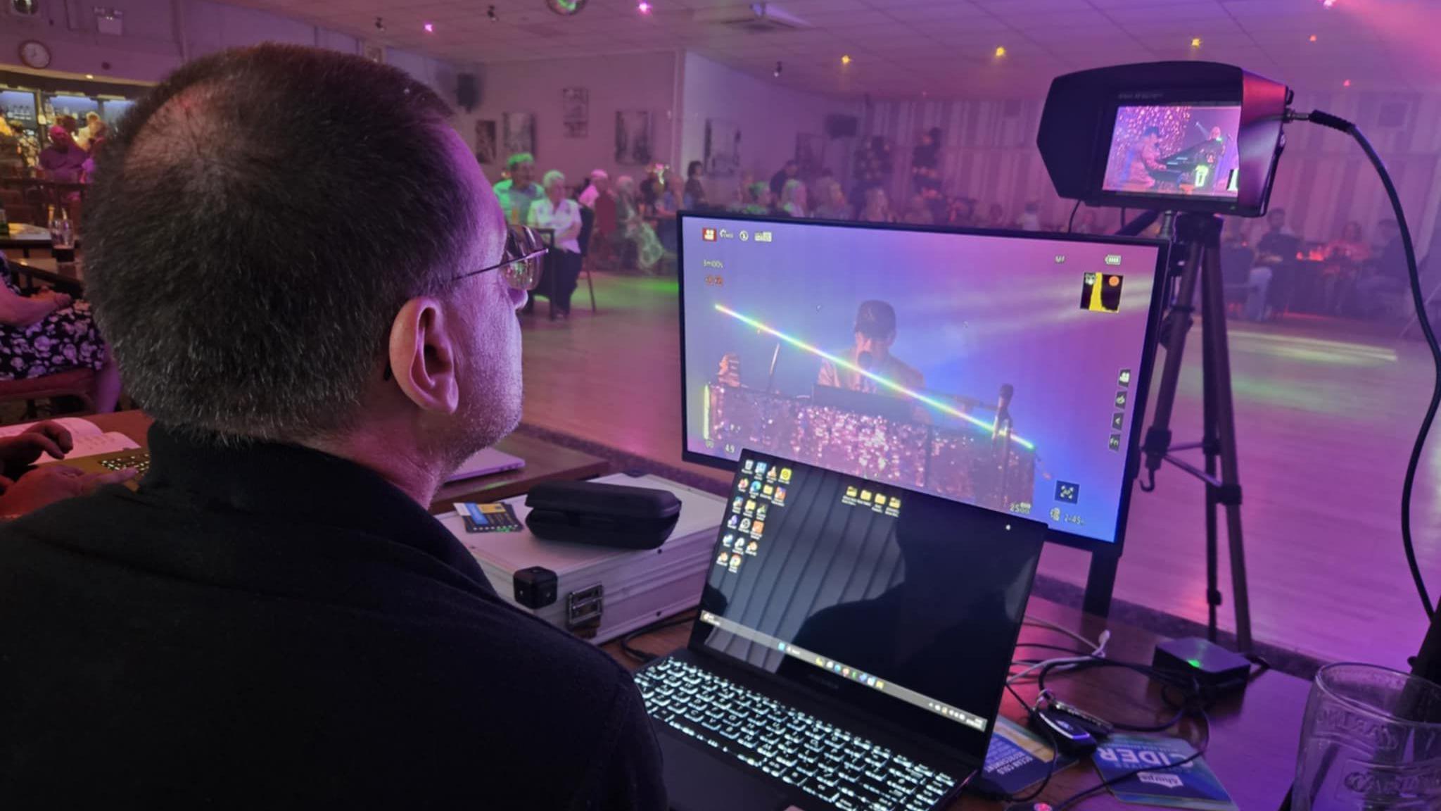 David Johnson looking at a small and a large screen as well as a camera as he films an indoor event where people are sitting around the perimeter of a dance floor.