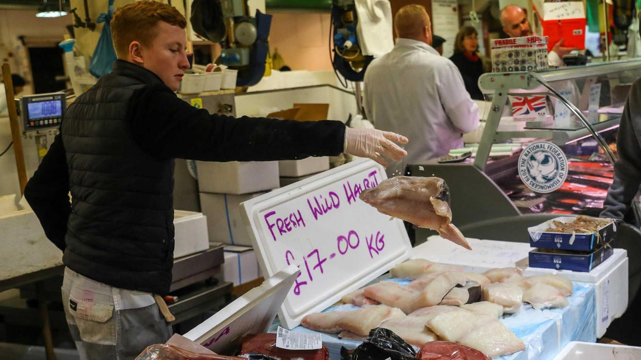 A fishnmonger tosses a halibut into pile of fish on a table in front of him. There is a sign that reads "fresh wild halibut, £17 per kg" behind the fish.  