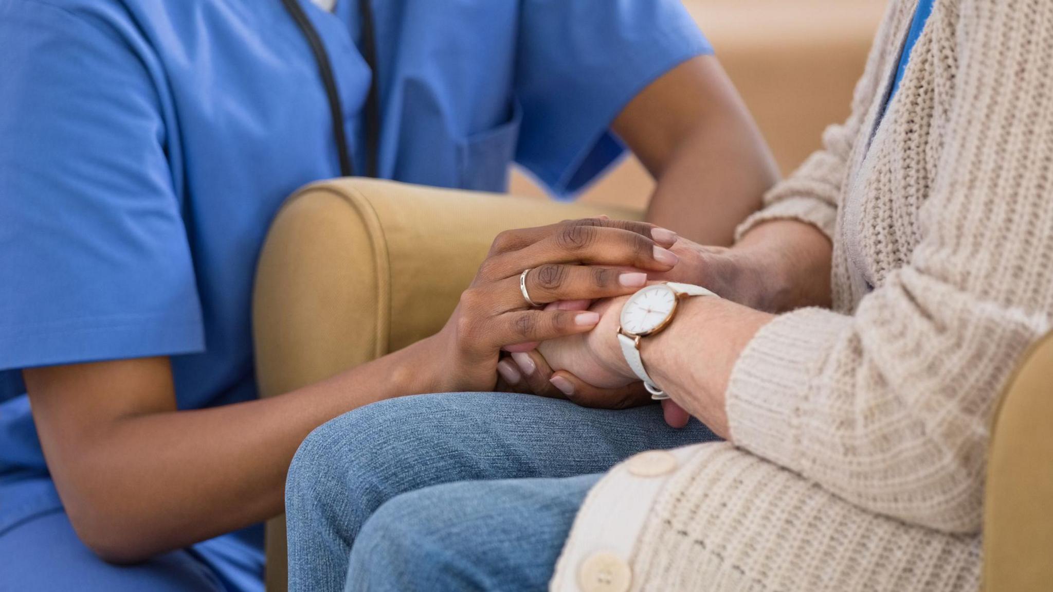 Healthcare professional in blue scrubs holding hands with a person wearing a beige knitted cardigan and jeans, seated on a light-coloured chair