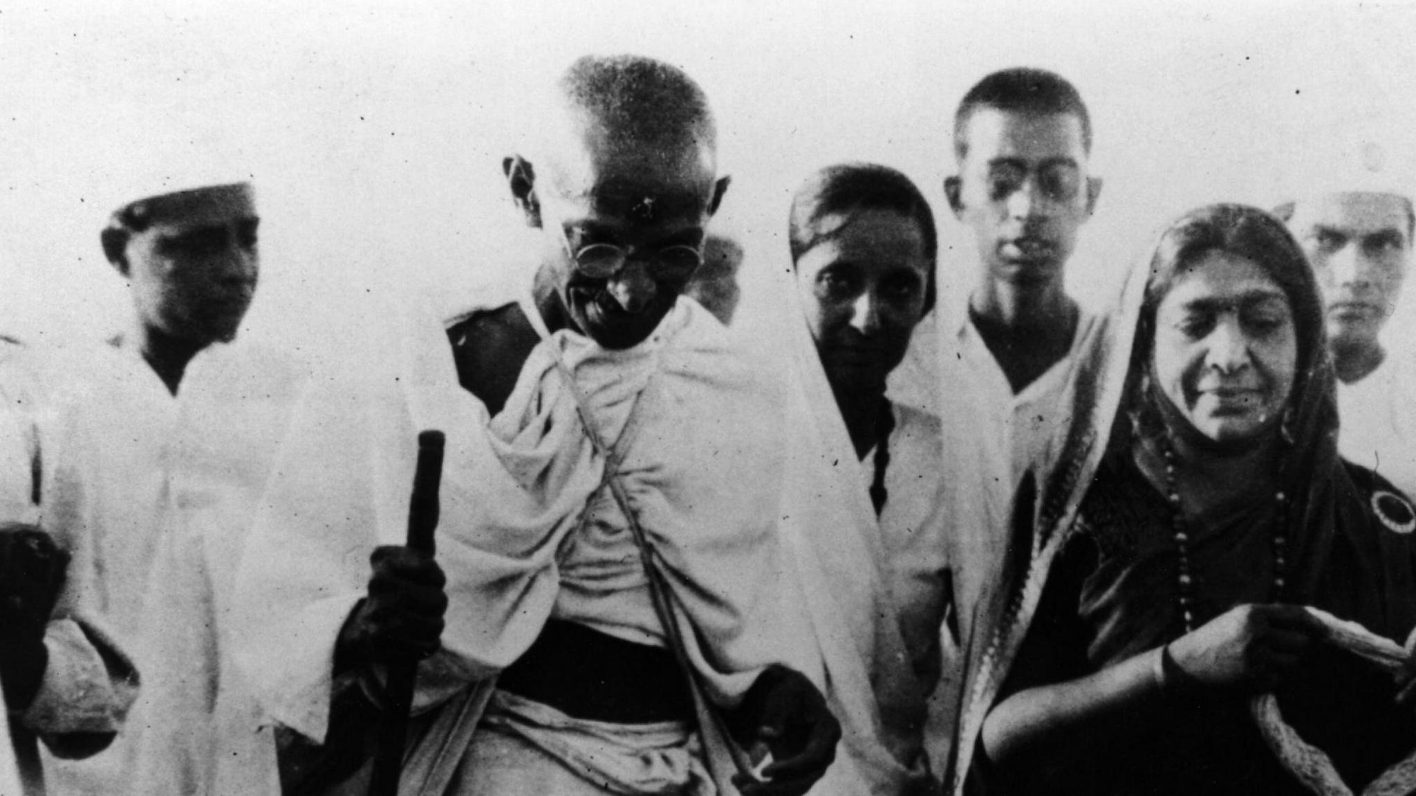 Mahatma Gandhi smiles and looks down as he strides with his staff at the front of a group of Indian men and women during the march in a black and white image.