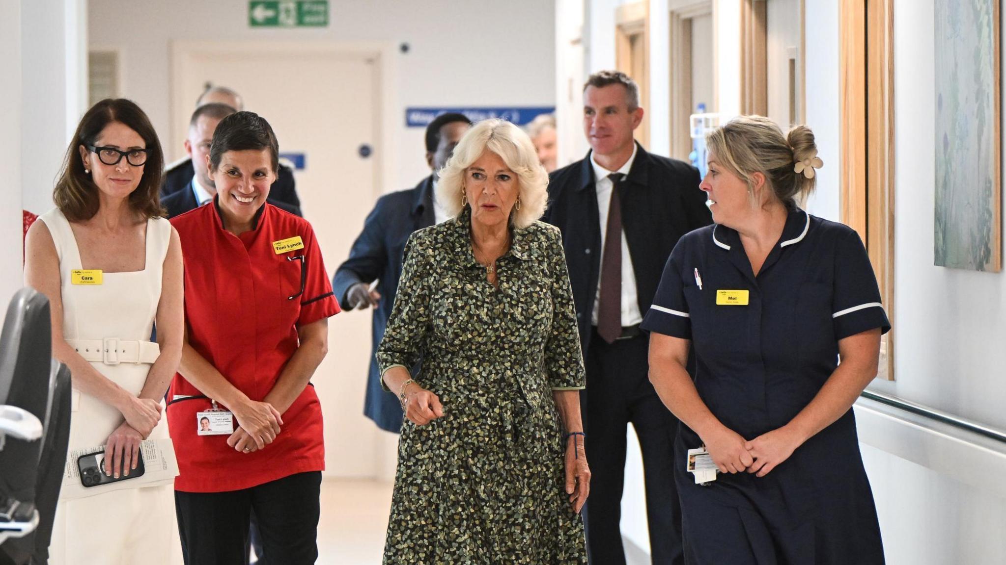 Cara Charles-Barks on the far left walking alongside two women dressed in NHS uniform, and Queen Camilla, during the opening of a new cancer centre. They are walking down a bright hospital corridor. Behind the women, four men in suits can also be seen.