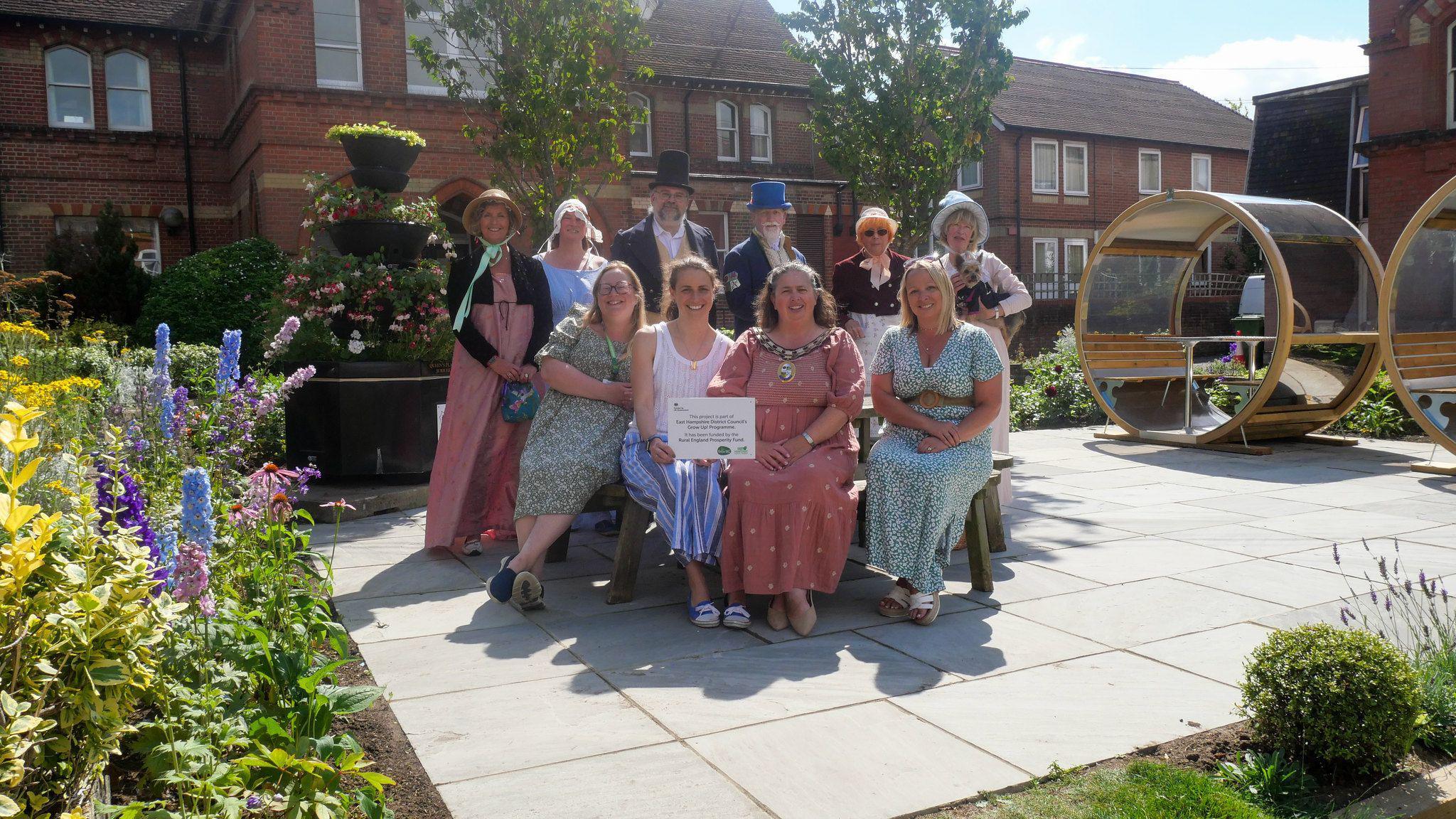 Alton Town Mayor, Councillor Annette Eyre, and EHDC local councillor, Ms Emily Young at the Jane Austen Memorial Garden with members of the Jane Austen Regency Week Committee. They are standing in the middle of the garden with flowers on the left and two benches on the right shaped like carriages.