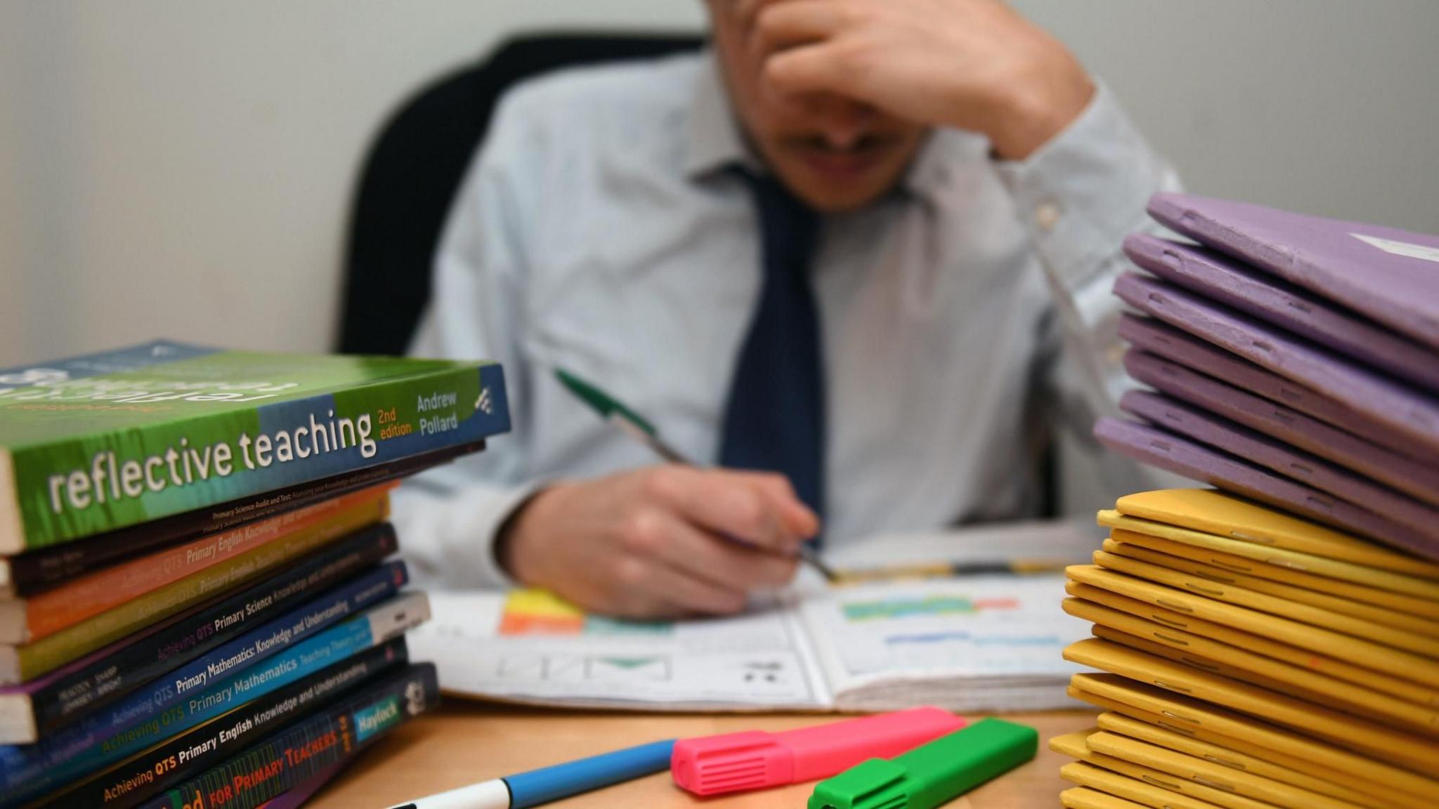 Stock image of a teacher marking at his desk. There are a pile of exercise books and reference books. The teacher's image is blurred