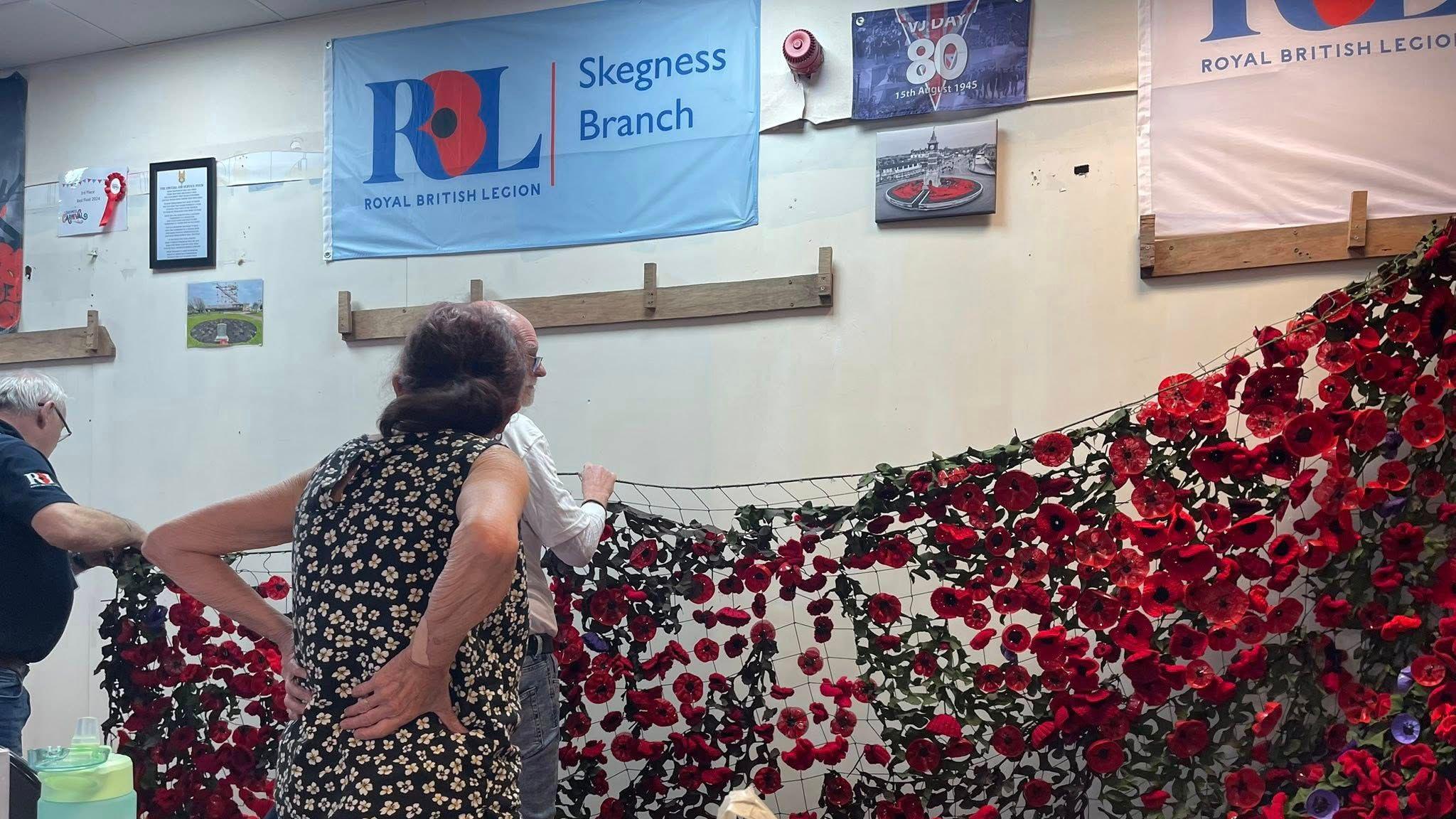 A net is being held by two men which is covered in handmade red knitted poppies. A woman can be seen standing in front of it with her hands on her back. A sign on the wall says 'RBL Skegness Branch'.