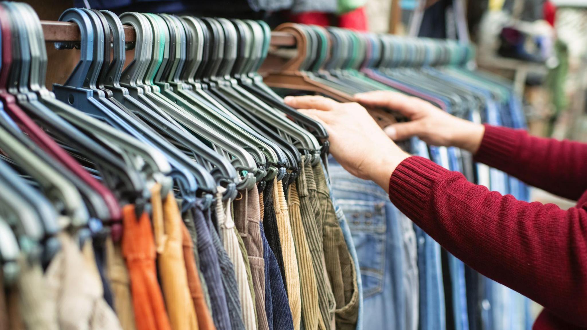 A bargain-hunter wearing a red jumper looking through a tightly packed rail of trousers on hangars