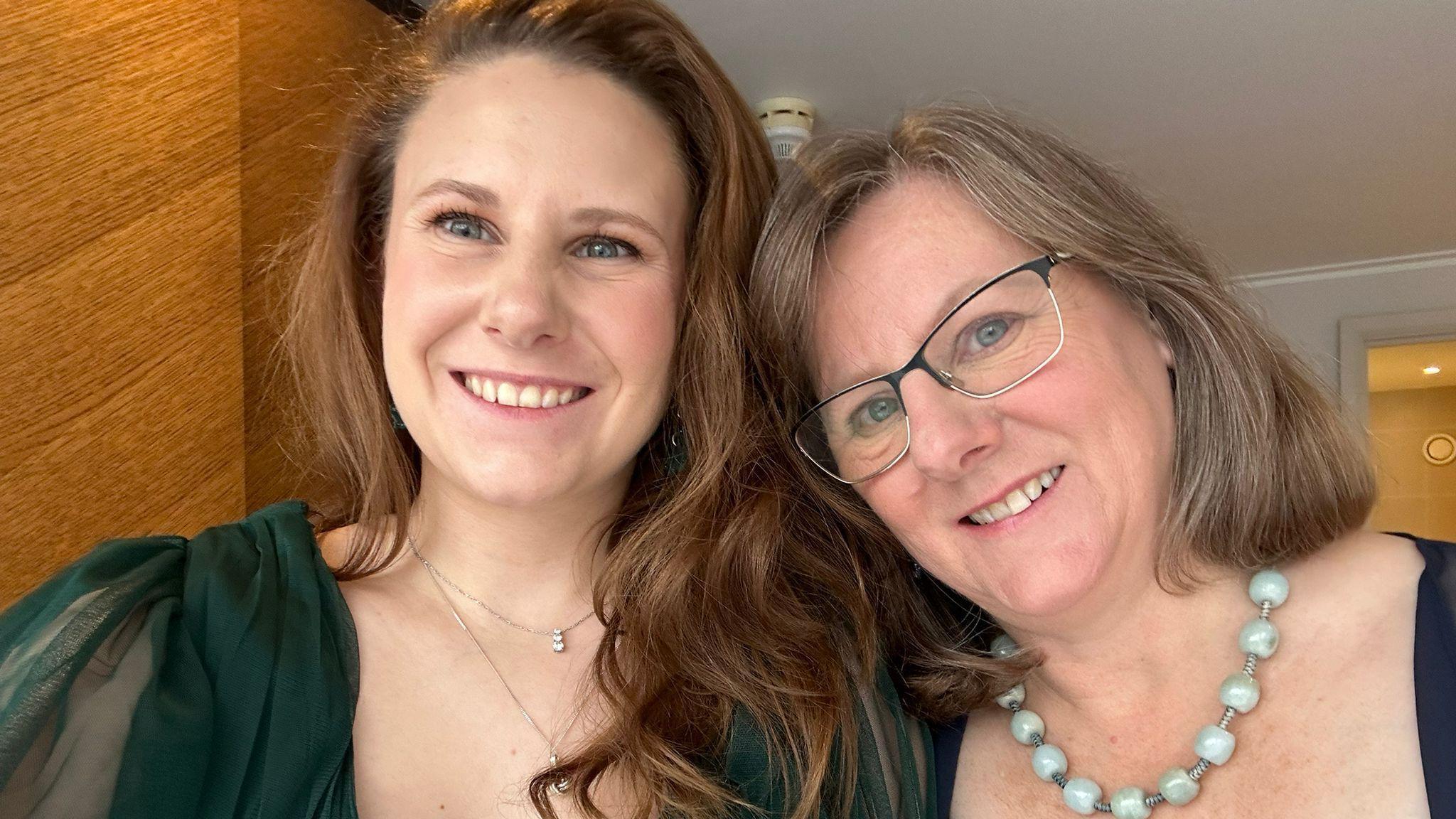 Molly and her mother smiling at the camera. Molly has long wavy hair and is wearing a green formal dress with two thin silver necklace chains. Her mother has shoulder length light brown hair and is wearing glasses and a chunky glass bead necklace. 