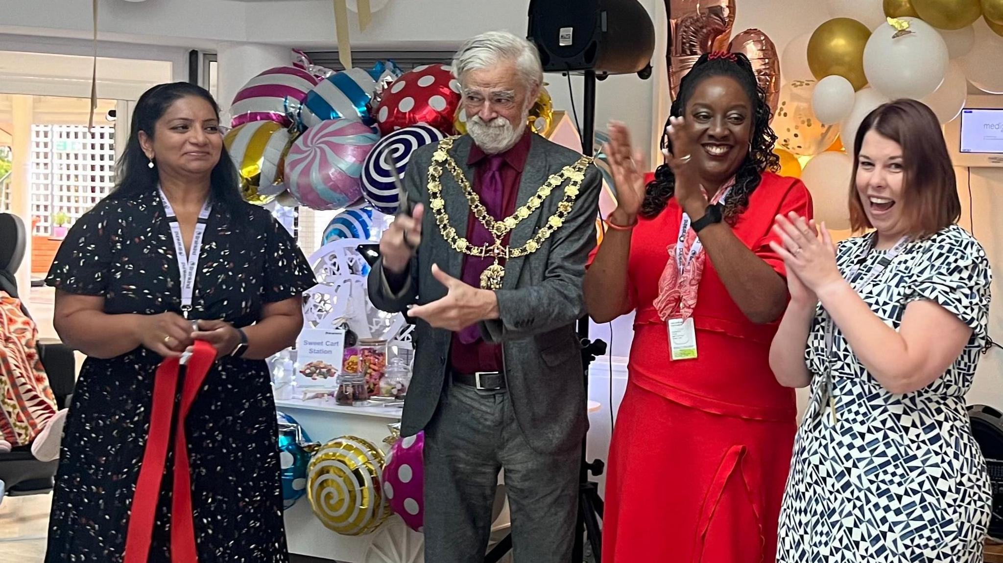 Alan Sherwell wearing a mayoral chain with Hazel Diaz-Fernandes holding a red ribbon, Irene Sobowale in a red dress and Claire Locke in a black and white dress clapping their hands at a party