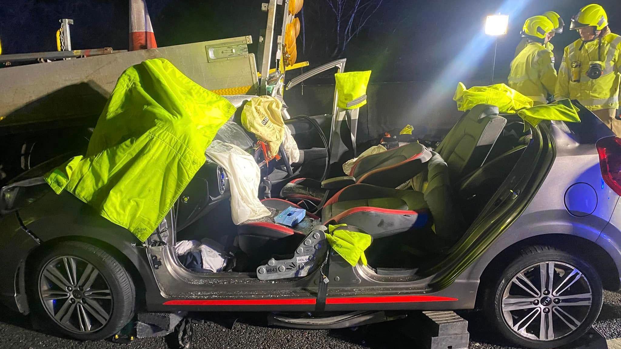 A closer vehicle of the car involved in the collision. Florescent yellow blankets have been placed over parts of it as firefighters talk nearby. The car is missing its roof, doors, front and back windscreen and windows.