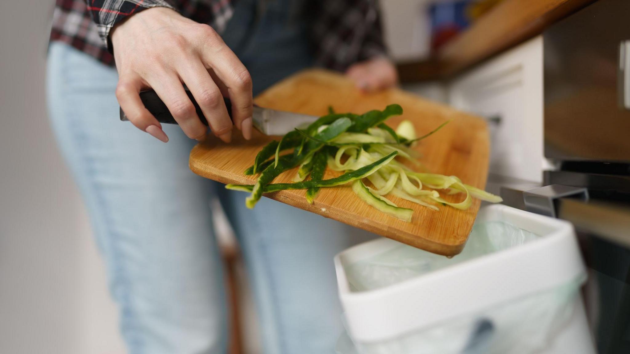 Close-up of vegetable peelings being scrapped from a wooden chopping board into a small white food caddy lined with a white bag. A woman's hands can be seen holding the chopping board and knife being used. 